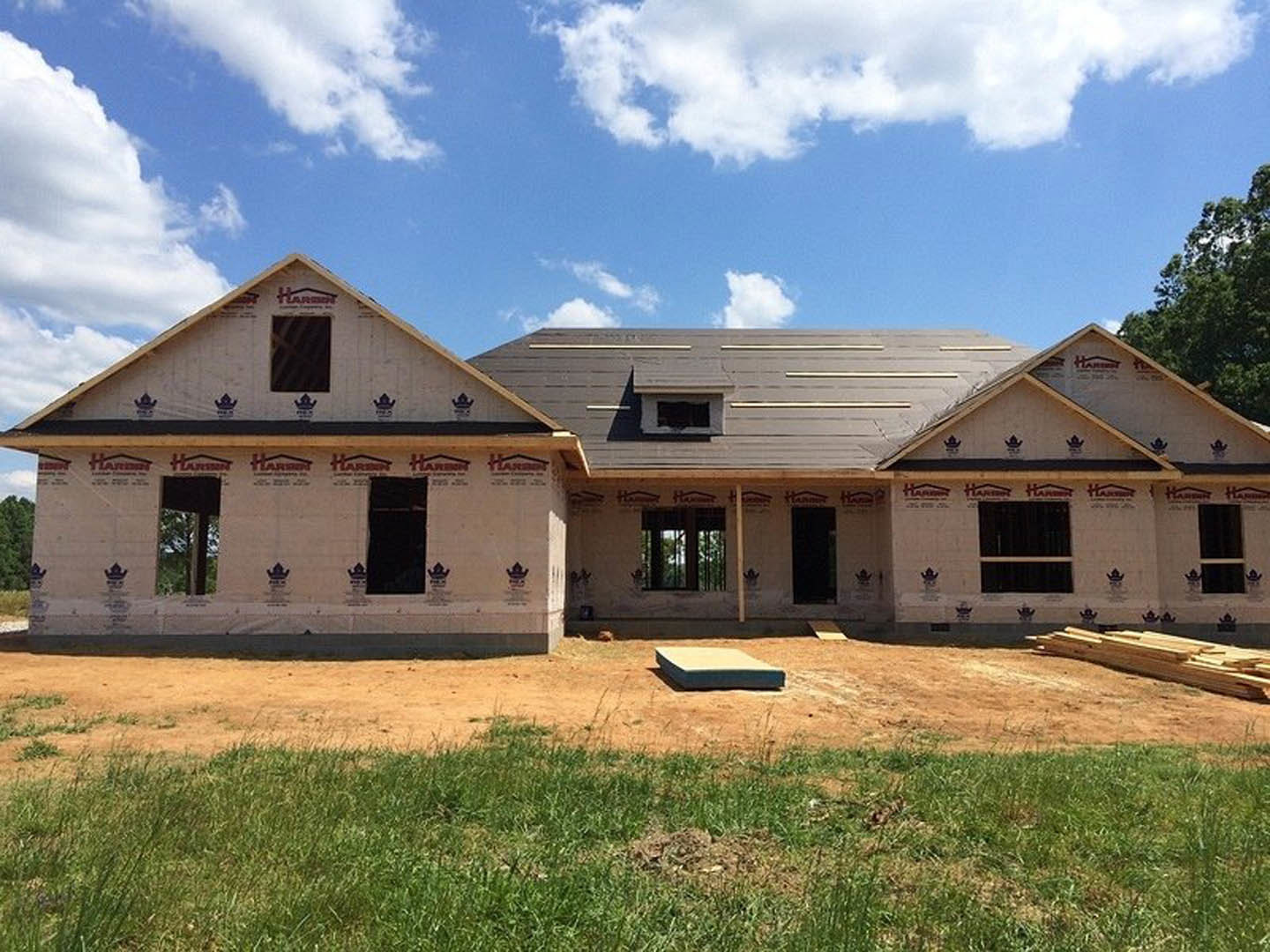 Partially built house with exposed framing and roof, surrounded by green grass under a blue sky, construction materials and dirt patch visible in foreground.