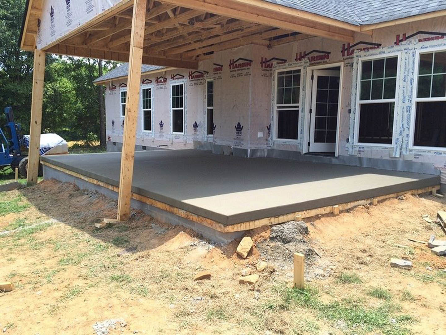 Wood-framed porch with exposed posts and beams atop a concrete foundation, unfinished exterior walls, window openings, and construction materials scattered on bare ground.
