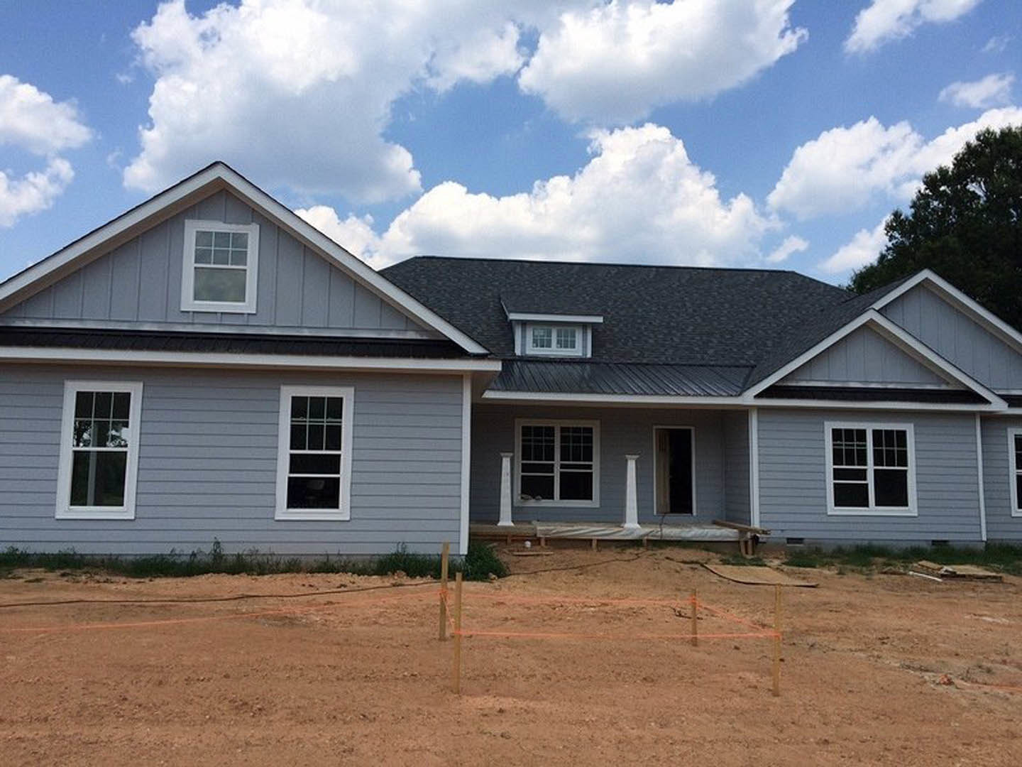 Two-story house under construction with exposed siding, white-framed windows, dirt foreground, orange rope fencing, and blue sky with scattered clouds