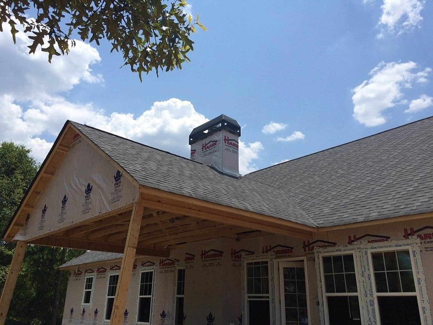 Partially built house with exposed framing, brick chimney, white sign on roof, square-paned window with white frame, tree branch in foreground, cloudy sky above