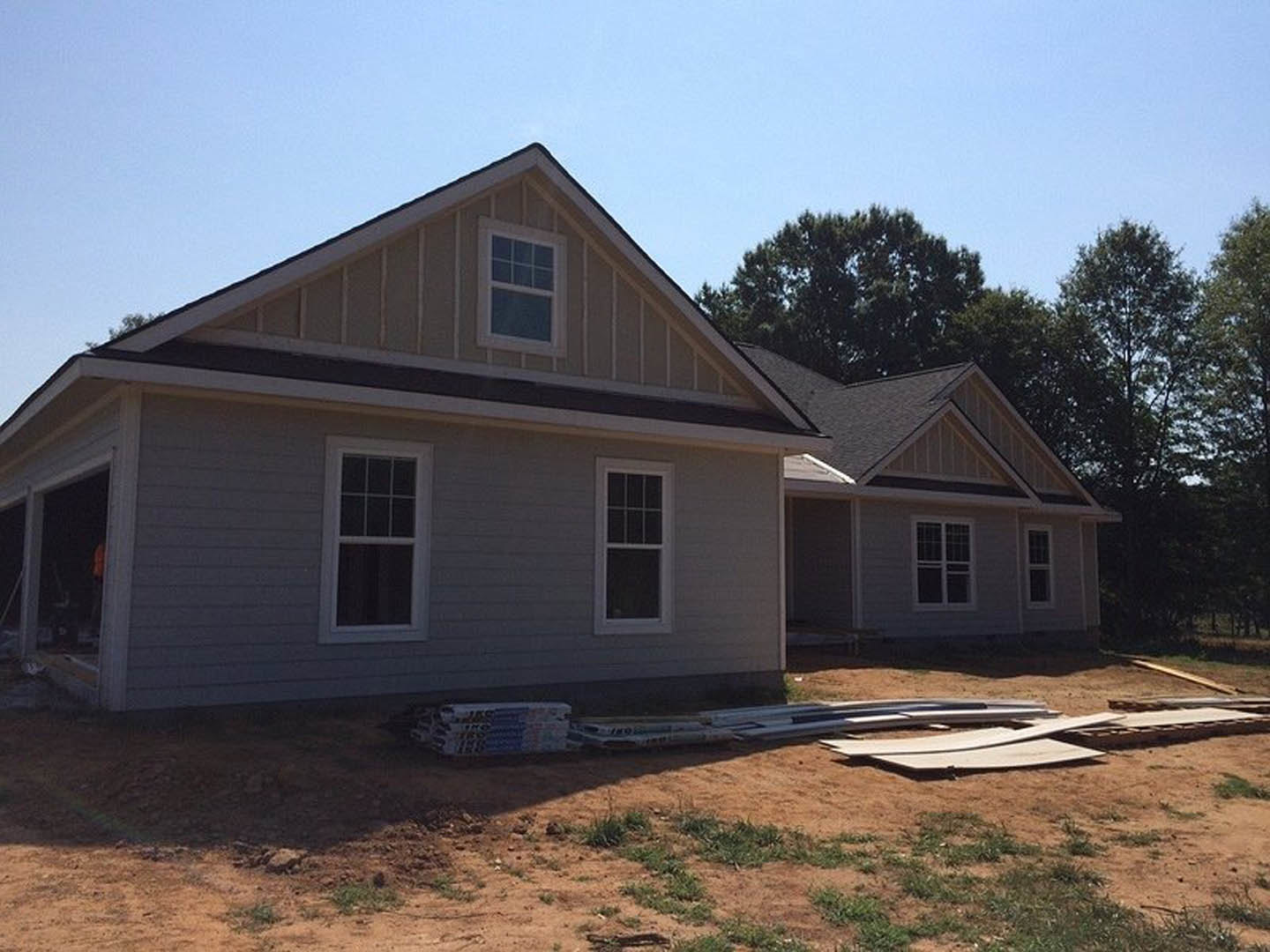 Partially built house with white framed multi-pane windows, gray siding, shingled roof, surrounded by grassy field and trees, American flag in foreground