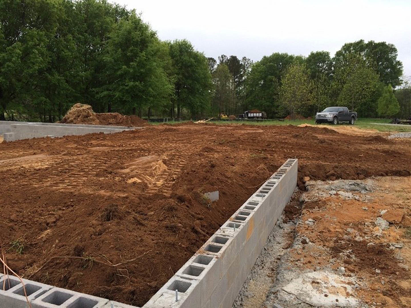 Concrete block foundation surrounded by dirt on a construction site, car parked on nearby road, group of trees in the background under open sky