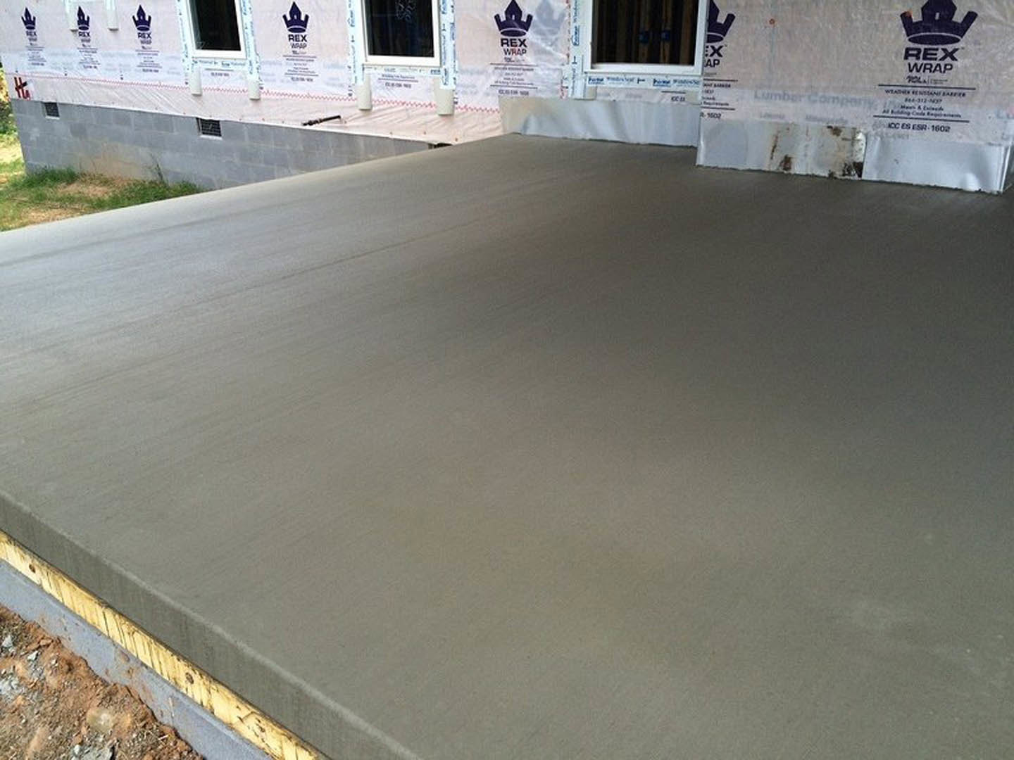 Smooth concrete slab foundation surrounded by grass, adjacent to a partially constructed building exterior