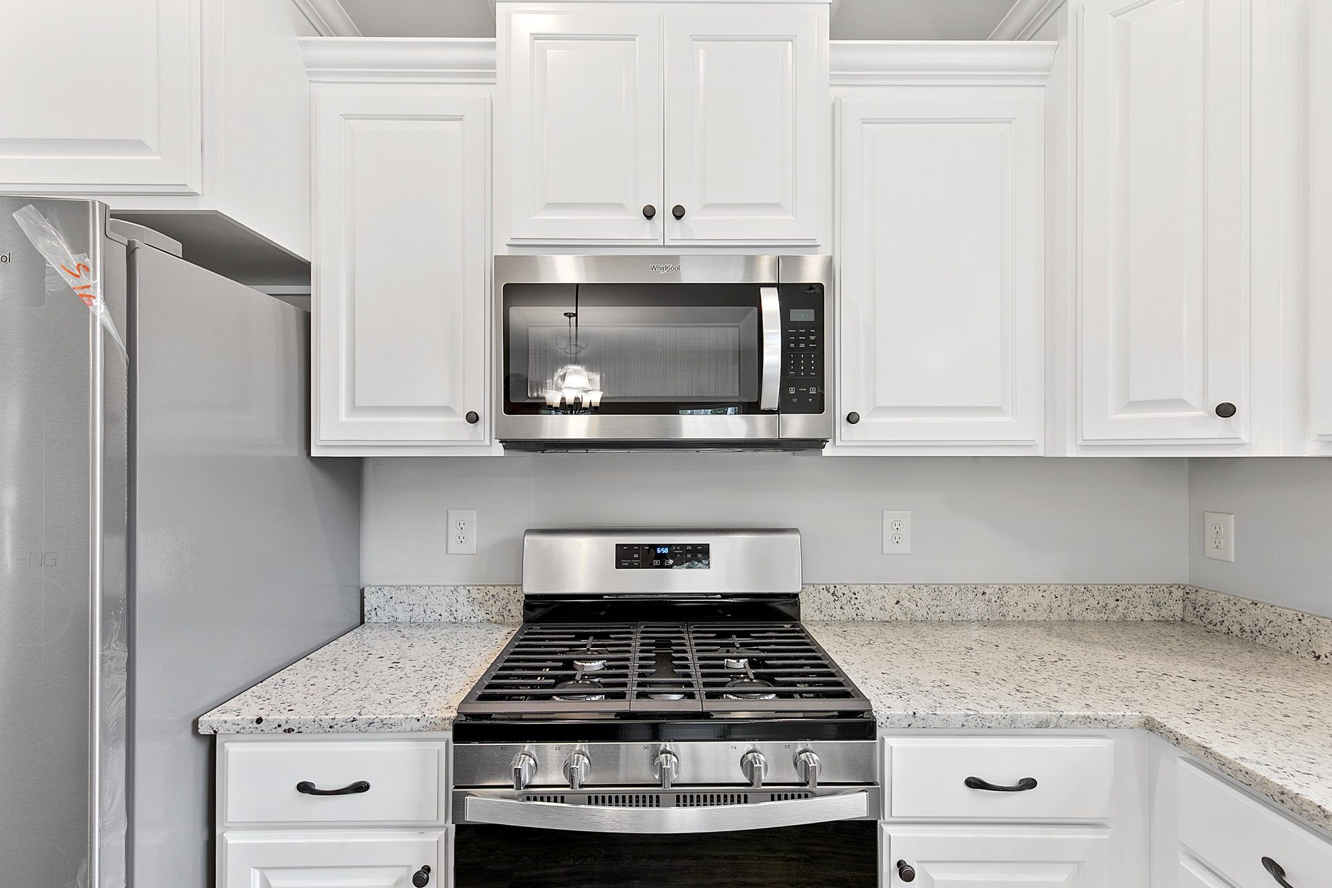 White kitchen cabinets, stainless steel microwave mounted above a stove with range and knobs, light countertops, silver cabinet handles, white door with shelving visible.