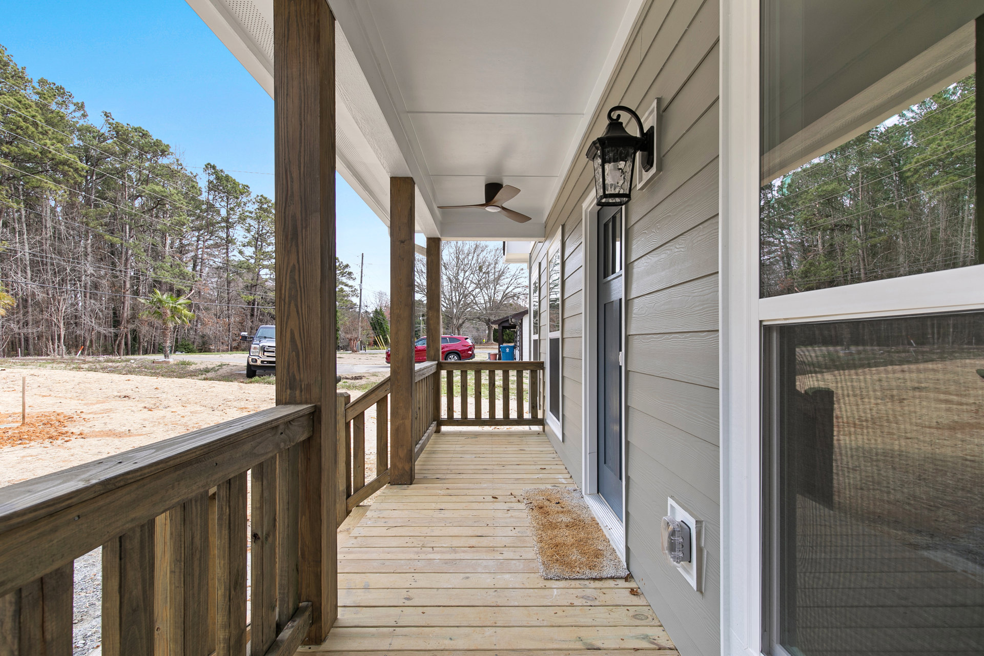Covered porch with wooden deck and railing, entry door, outdoor lamp, rug on wood floor, black truck and red car parked near wooden post, trees and sky in background