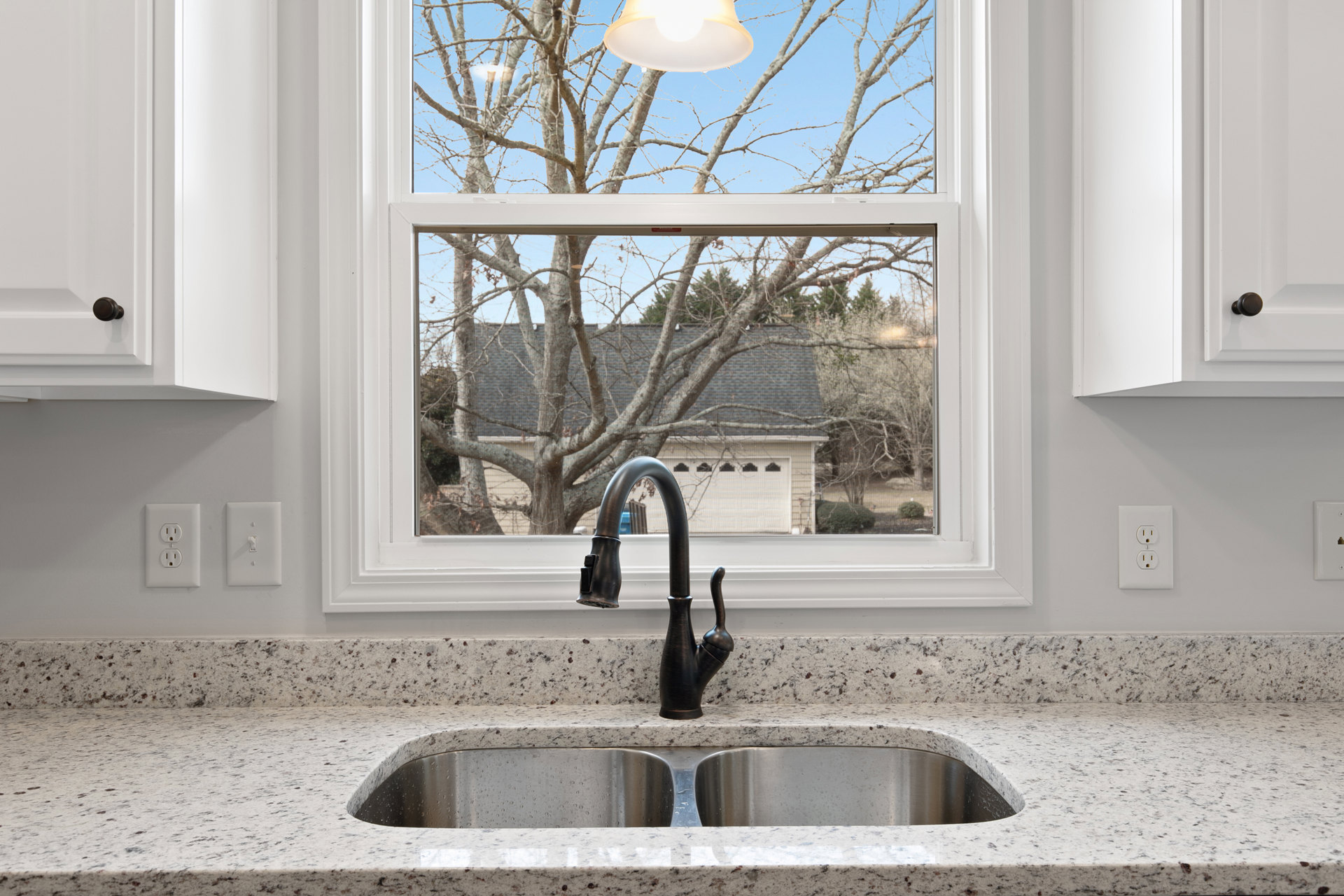 Stainless steel undermount sink with modern chrome faucet set in quartz countertop beneath a large window, white subway tile backsplash, and nearby electrical outlet