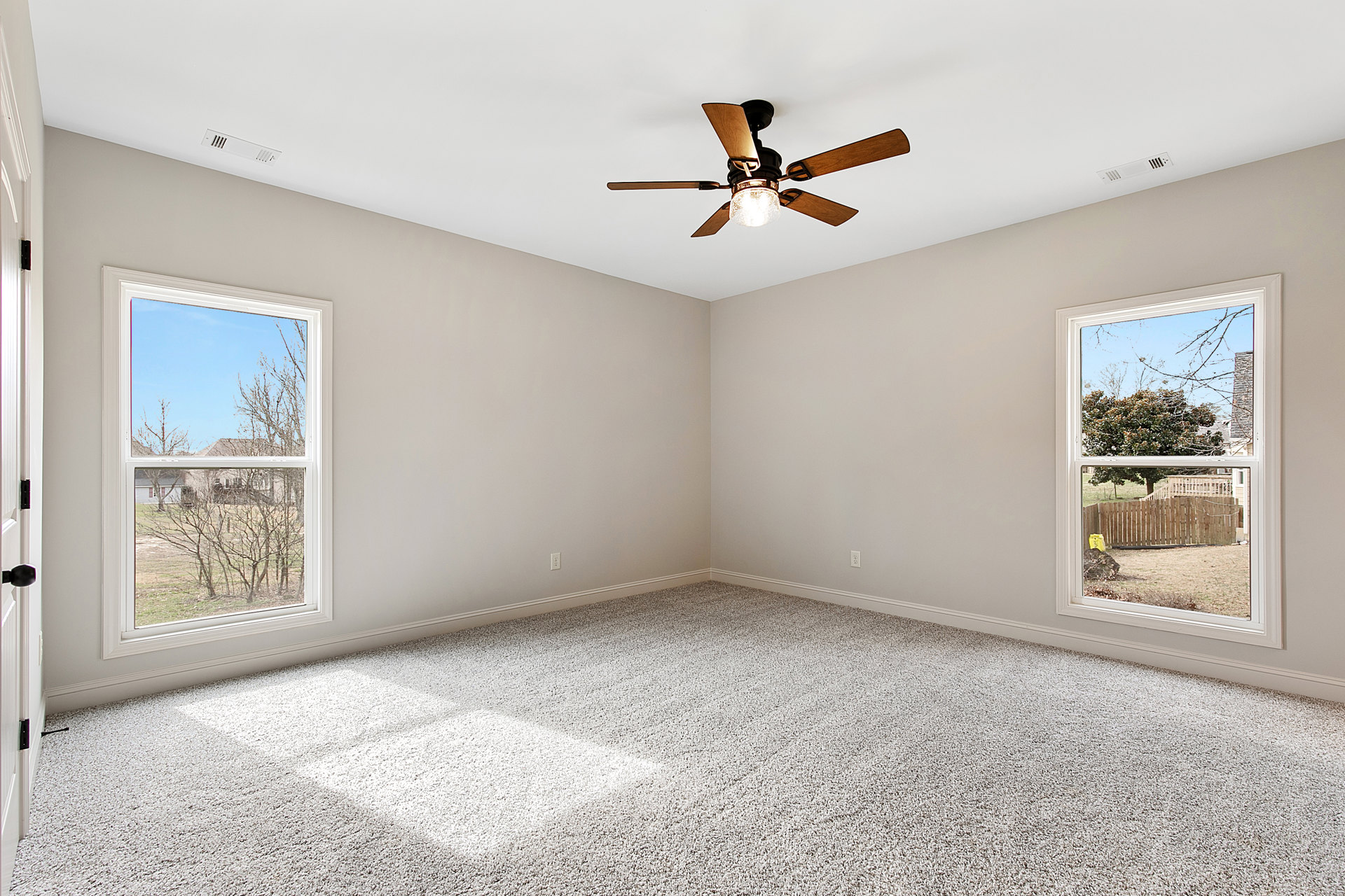 Carpeted room with white plaster walls, ceiling fan with light fixture, two windows showing trees and a wooden fence outside