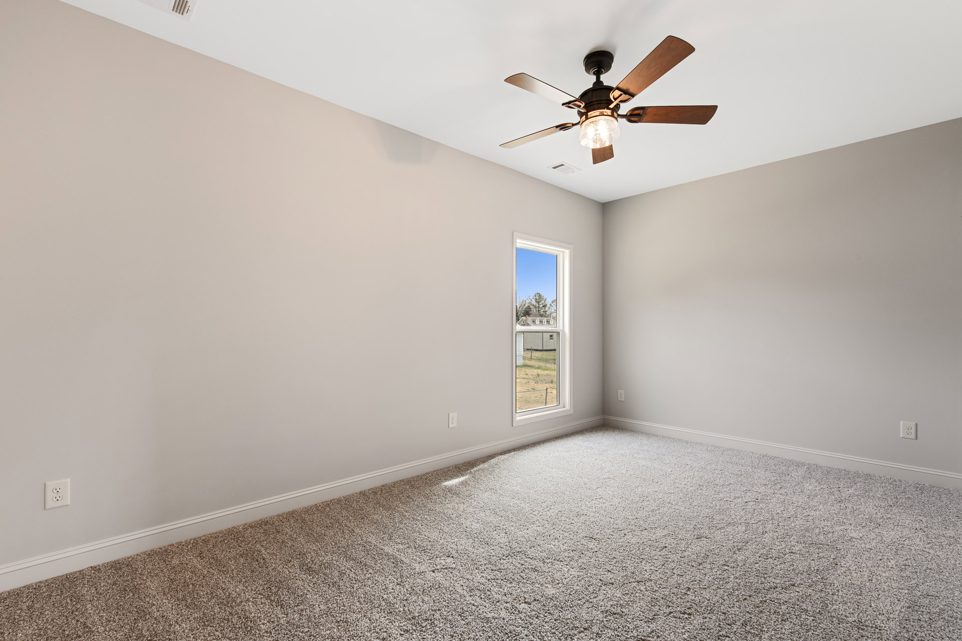 Neutral-toned carpeted room with white plaster walls, ceiling fan with integrated light, electrical outlet, and window overlooking neighboring house and trees