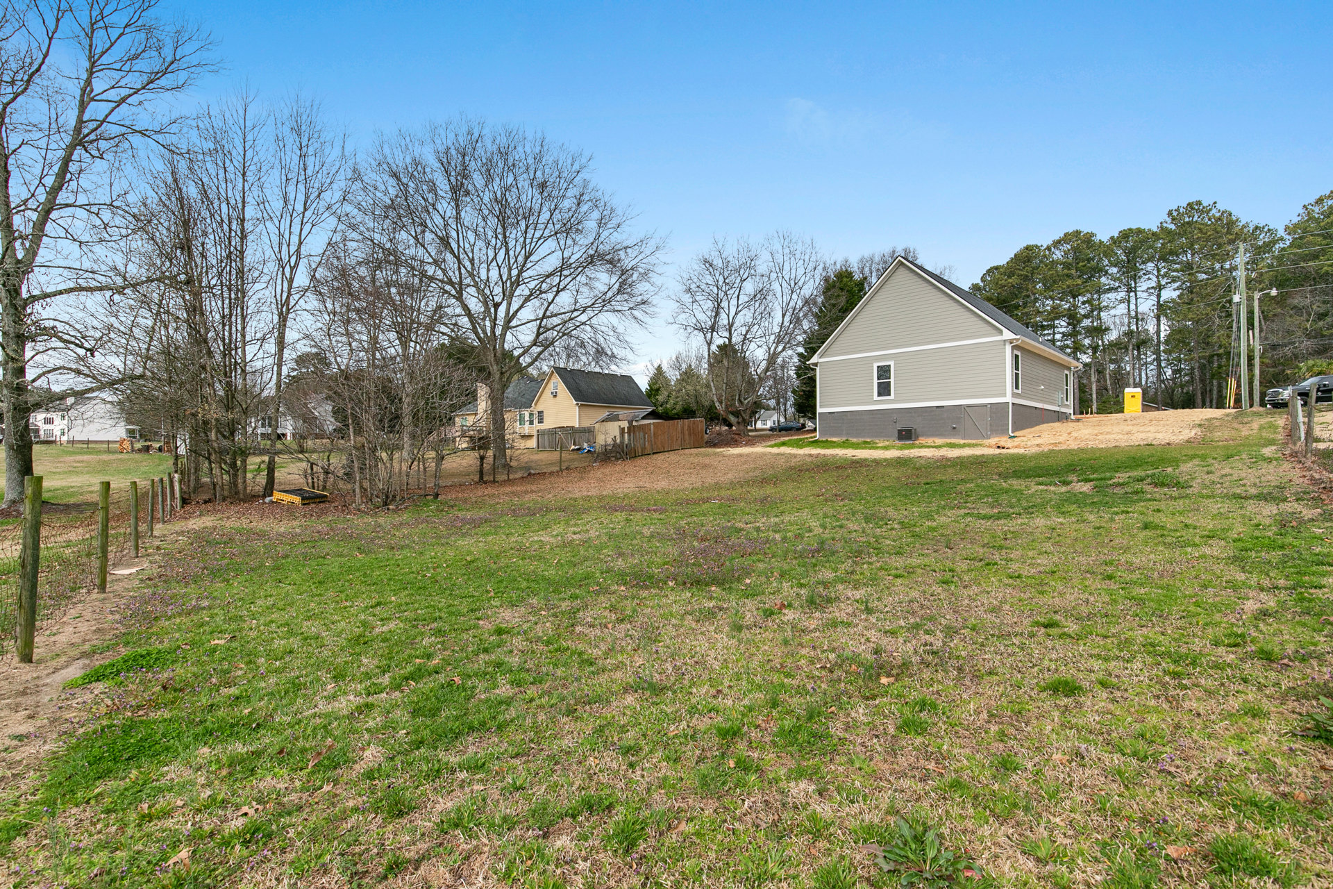 Two-story farmhouse with white siding, gabled roof, attached garage, wooden fence, mature trees, and manicured grass lawn under a clear blue sky
