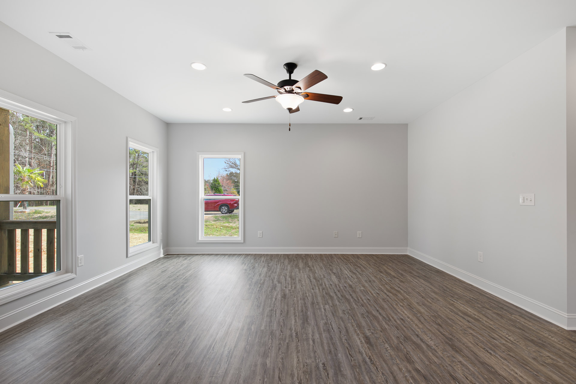 Wood flooring in a room with plaster walls, ceiling fan with light fixture, large window overlooking a red car parked outside and wooden fence.