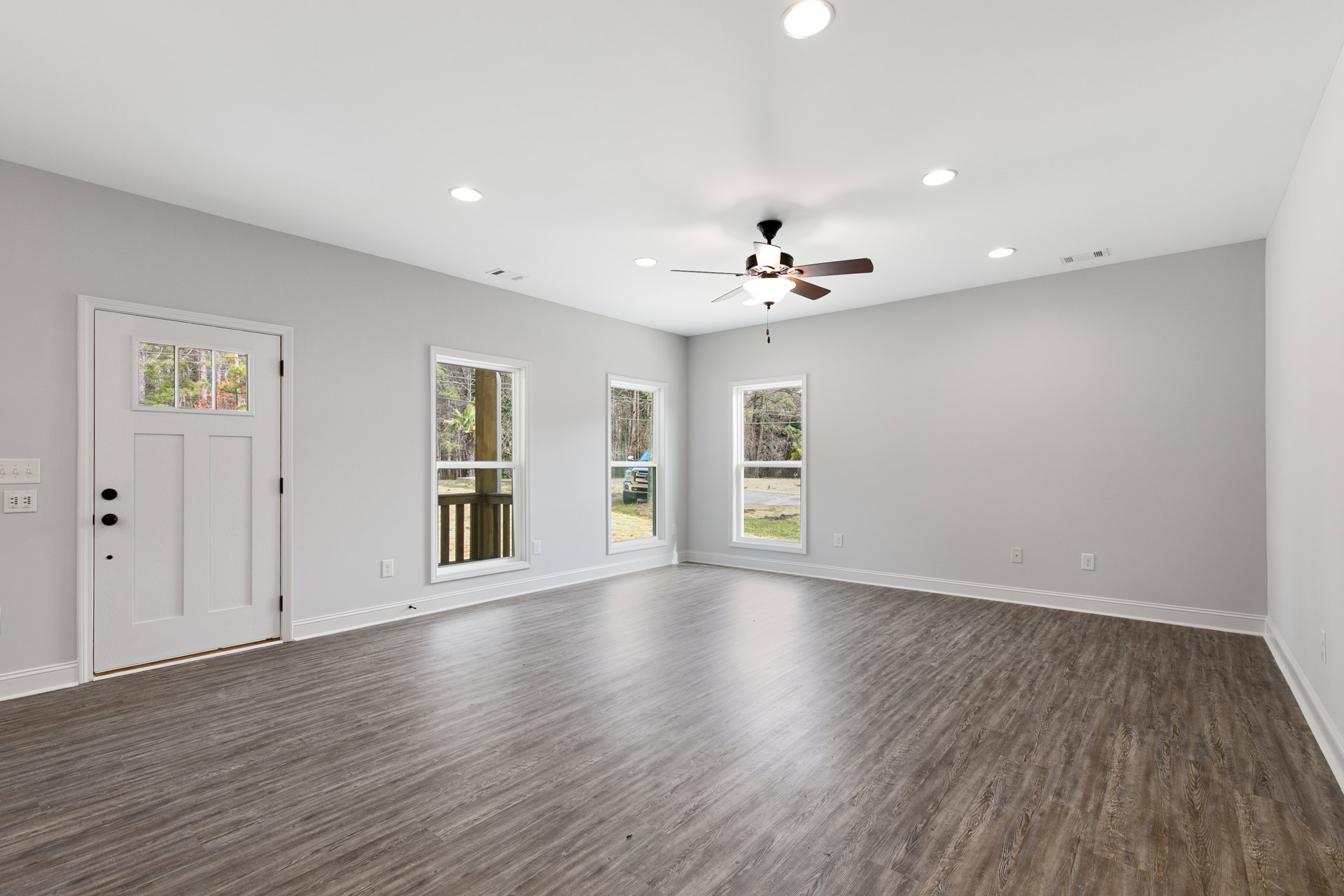 Ceiling fan with light fixture, white walls, laminate wood flooring, multiple windows showing trees and grass, white door with glass inset