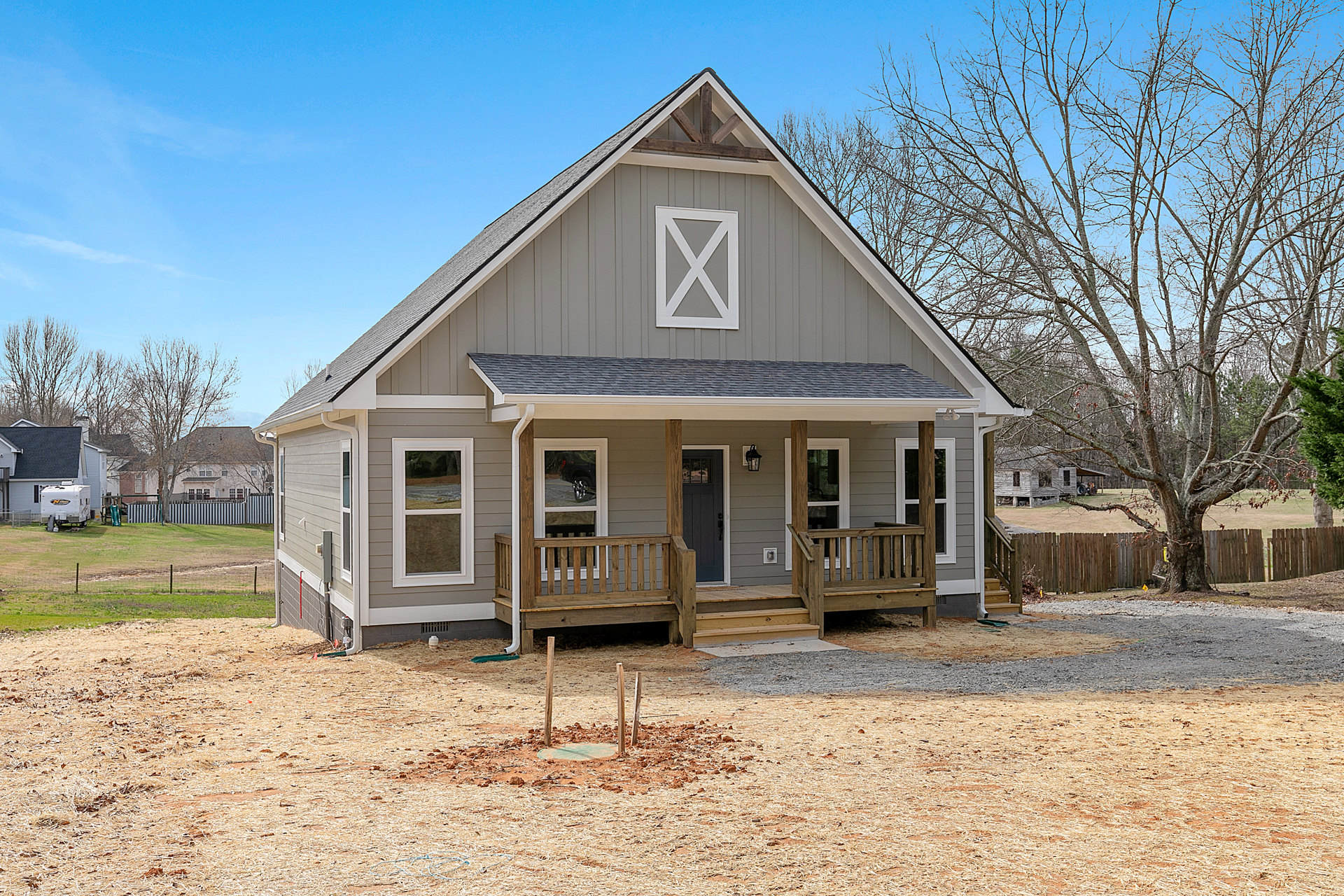 Gray-sided house with covered porch, large front yard, mature trees in background, prominent windows, and shingled roof.