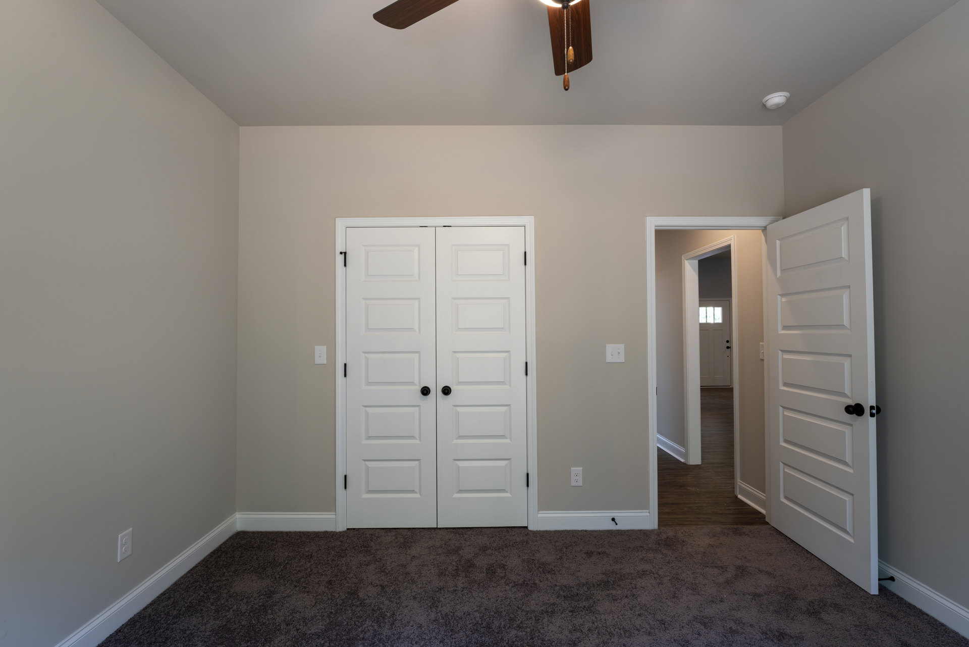 Neutral-toned carpeted room with white double doors featuring black knobs, single white door with black knob, and ceiling fan mounted on smooth plaster ceiling.