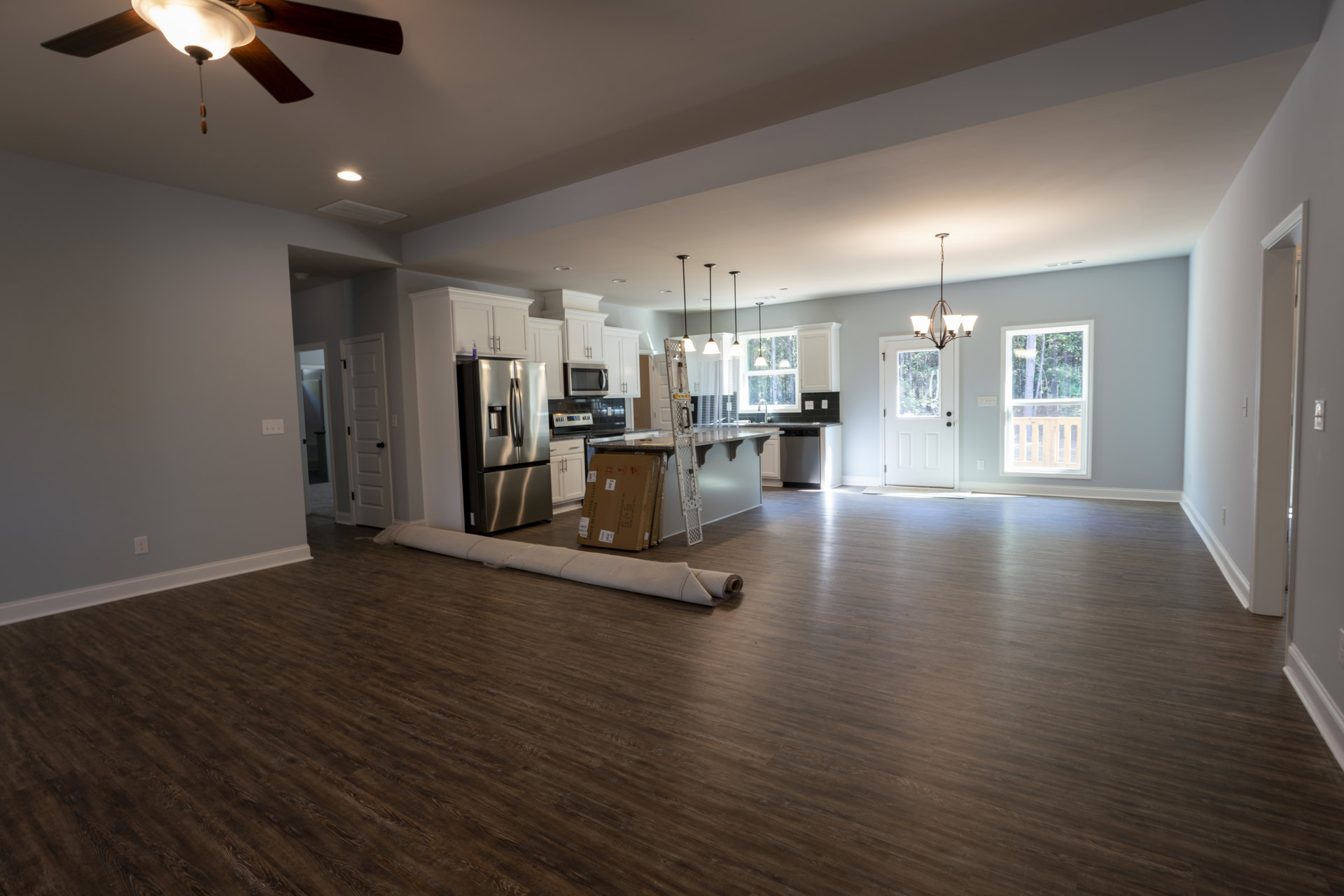 Open kitchen with stainless steel double-door refrigerator, large wooden dining table, ceiling fan with light, roll of fabric on table, carpet roll on laminate wood flooring