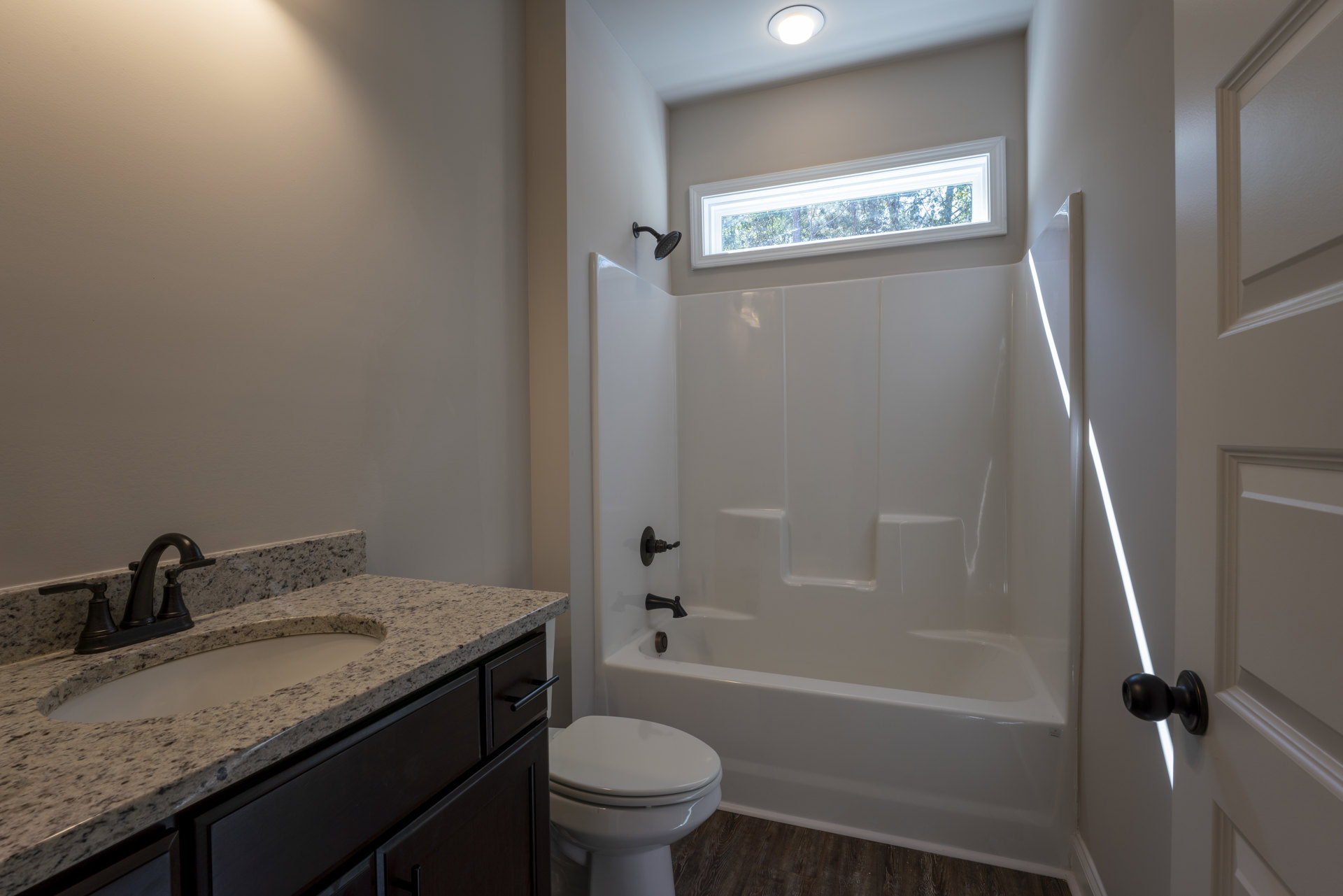 Bathroom with white toilet, rectangular sink set in a wood vanity, wall-mounted mirror, ceiling light fixture, window showing green trees, and white bathtub with tiled walls.