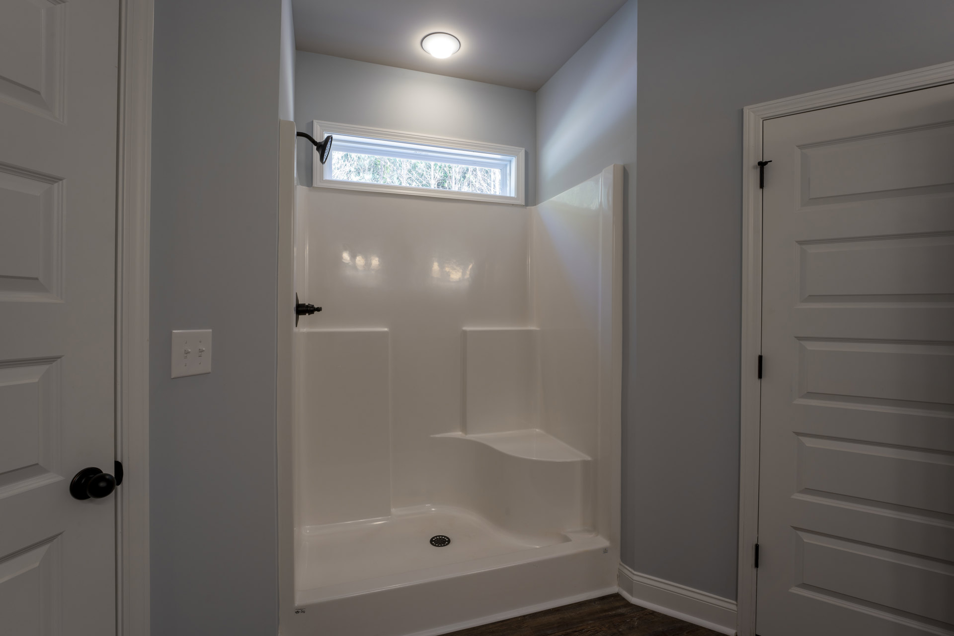 Glass-enclosed shower with ceiling light, white door featuring black hinge, window overlooking trees, and light switch on pale wall