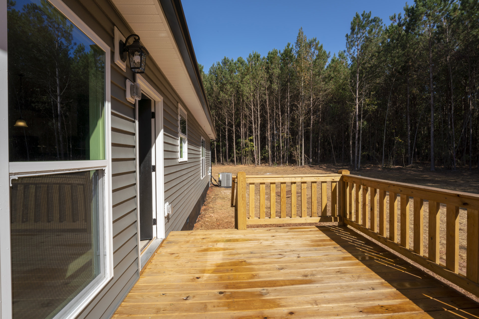 Wooden deck with railing, surrounded by mature trees and blue sky, house exterior features window with screen and wooden fence in background