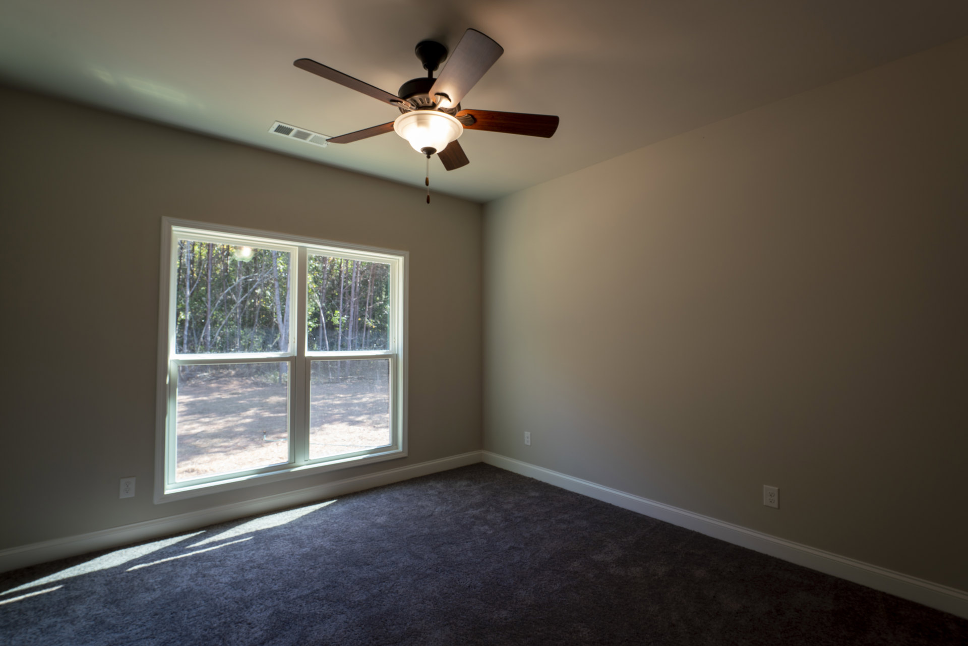 Neutral-toned room with carpet flooring, ceiling fan with light fixture, large window showing leafy trees outside, white plaster walls
