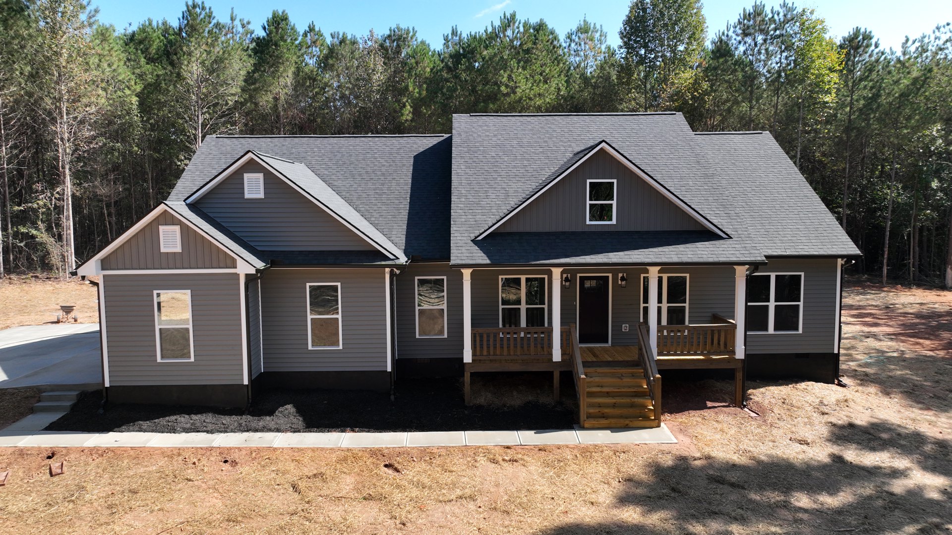 Grey siding house with white trim, covered front porch, concrete driveway, multiple windows, mature trees in the background, blue sky overhead.