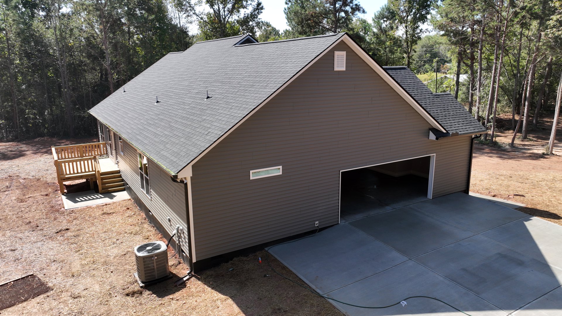 Modern house exterior with attached garage, concrete driveway, wooden bench on deck, air conditioner unit beside wall, large window, shingled roof, and mature trees in background