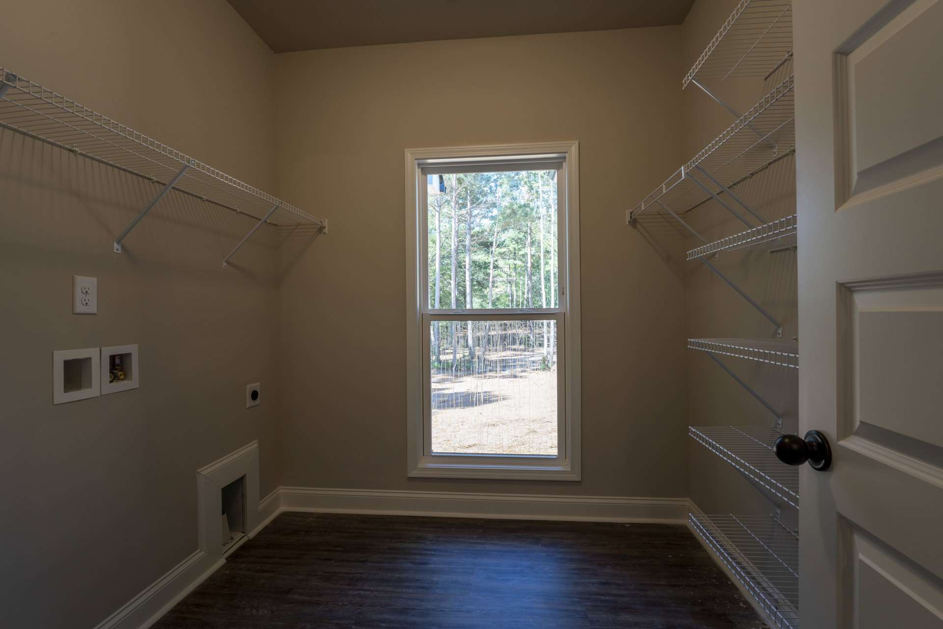 Built-in white shelves beside a large window with trees visible outside, dark wood flooring, white baseboards, electrical outlet, black balloon tied to a door handle