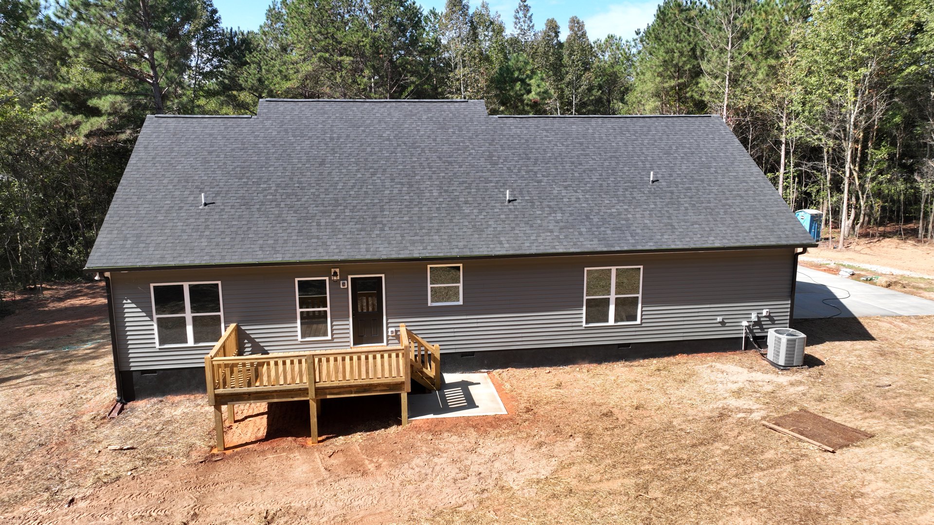 Wooden deck attached to a light-colored house, black door with glass window, white-framed windows, white heat pump unit beside the exterior, grassy yard bordered by trees