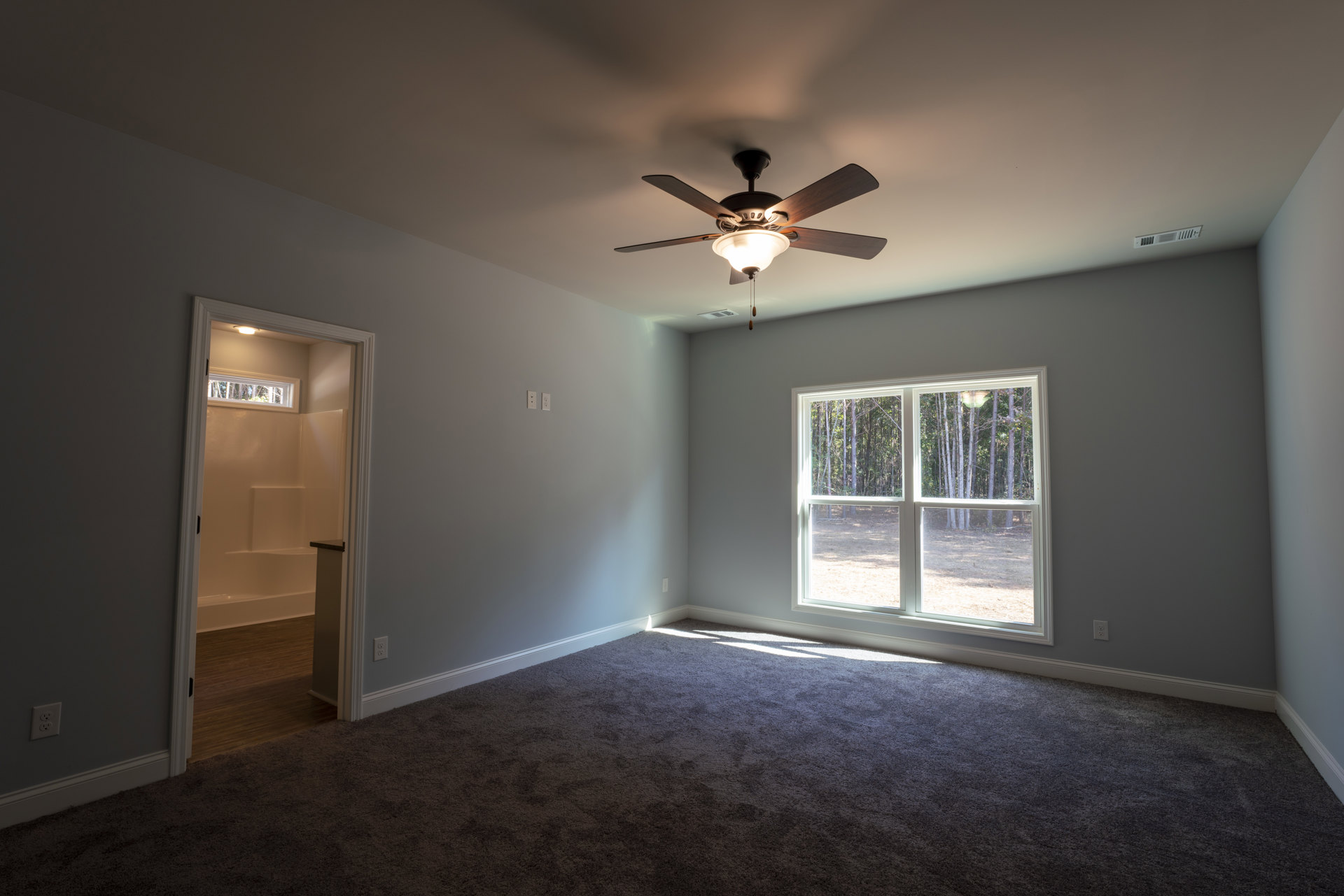 Neutral-toned room with plaster walls, ceiling fan with integrated light fixture, large window revealing leafy trees outside, hardwood flooring