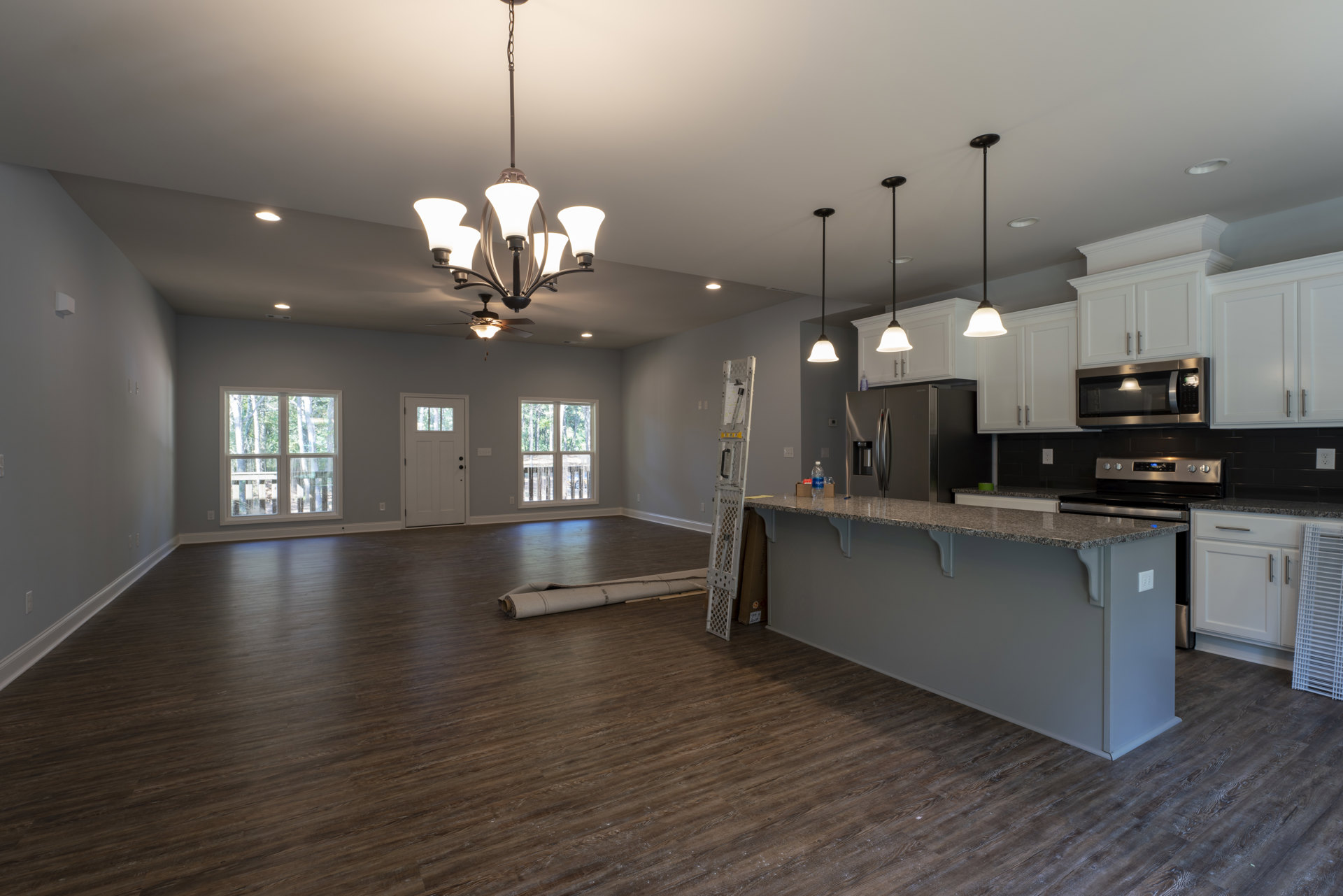 Open kitchen and living room with wood flooring, white cabinetry, stone countertops, stainless steel appliances, window with tree view, white framed window and door, ceiling