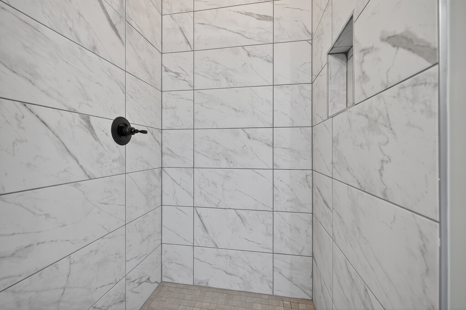 White tile shower with black shower head and handles, stone tile floor, square window on tile wall