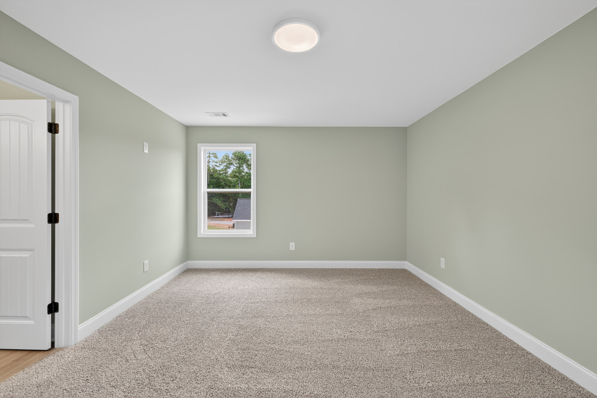 Bedroom with beige carpet, white walls, large window overlooking trees, recessed ceiling light, and white door with visible hinge