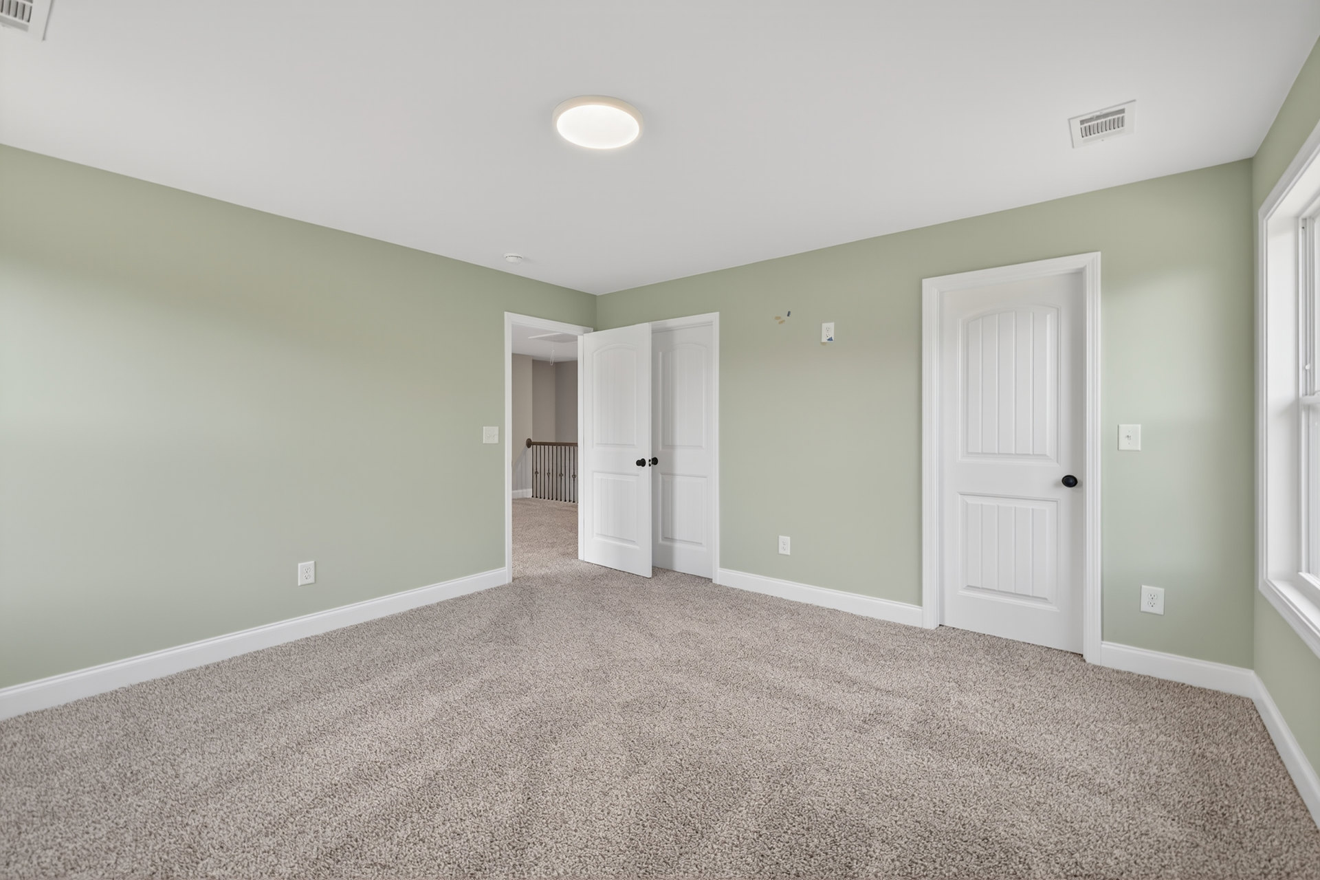 Green-painted walls, white paneled doors with black handles, white ceiling vent, carpeted floor, white baseboards, close-up of modern light fixture and stair railing