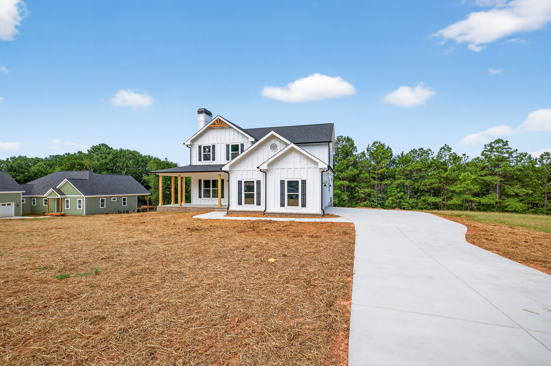 White house with black shutters and white roof, front porch, curved concrete driveway, green lawn, mature trees in background, cloudy sky
