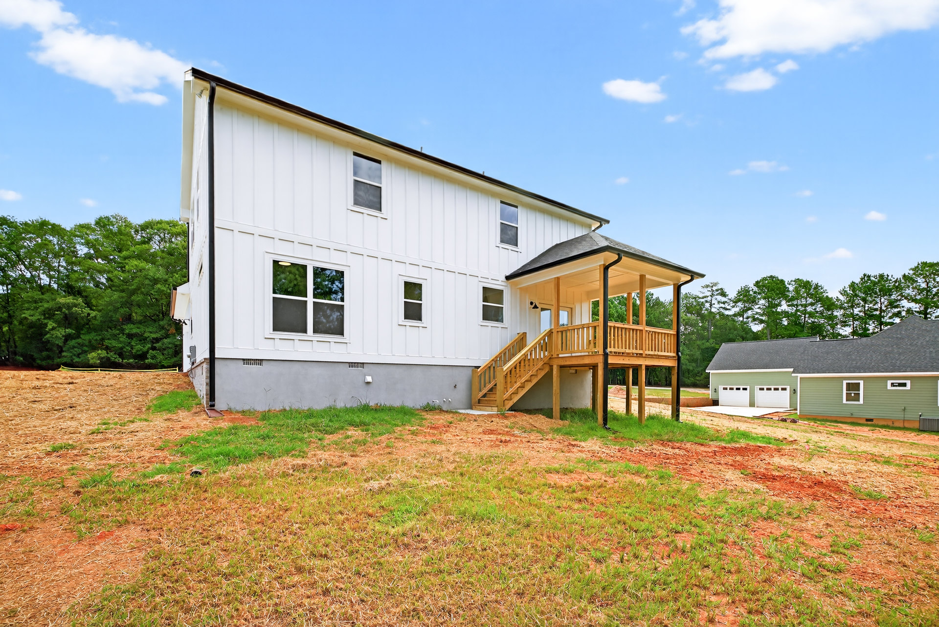 White house with attached garage, wooden porch featuring wood railing and staircase, landscaped front yard with grass and trees, large windows, cloudy sky overhead