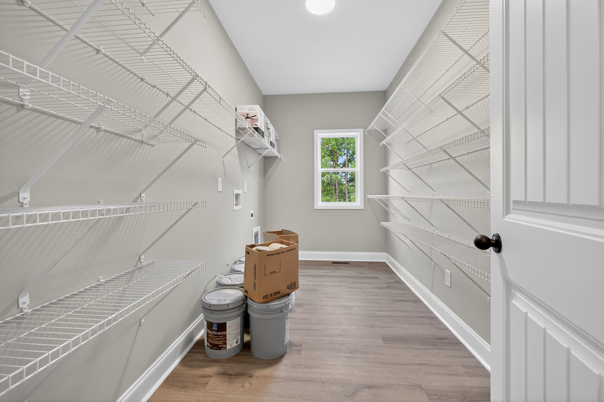 White built-in shelves holding plastic buckets and cardboard boxes, window with trees visible outside, light plaster walls, wood flooring, and a close-up of a silver door knob.