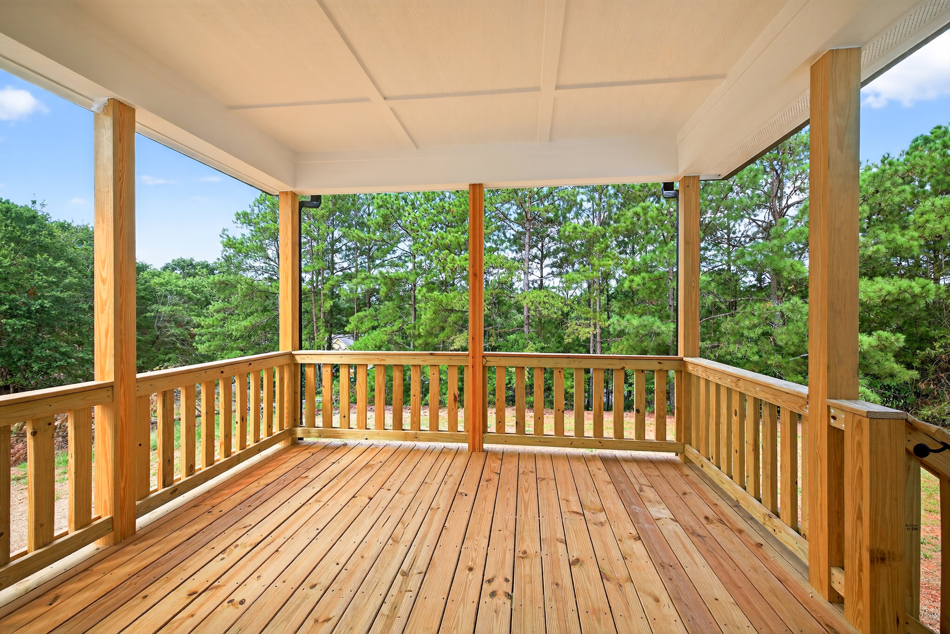 Wooden deck with railing, white wood-paneled ceiling, large green trees in background, shaded outdoor space
