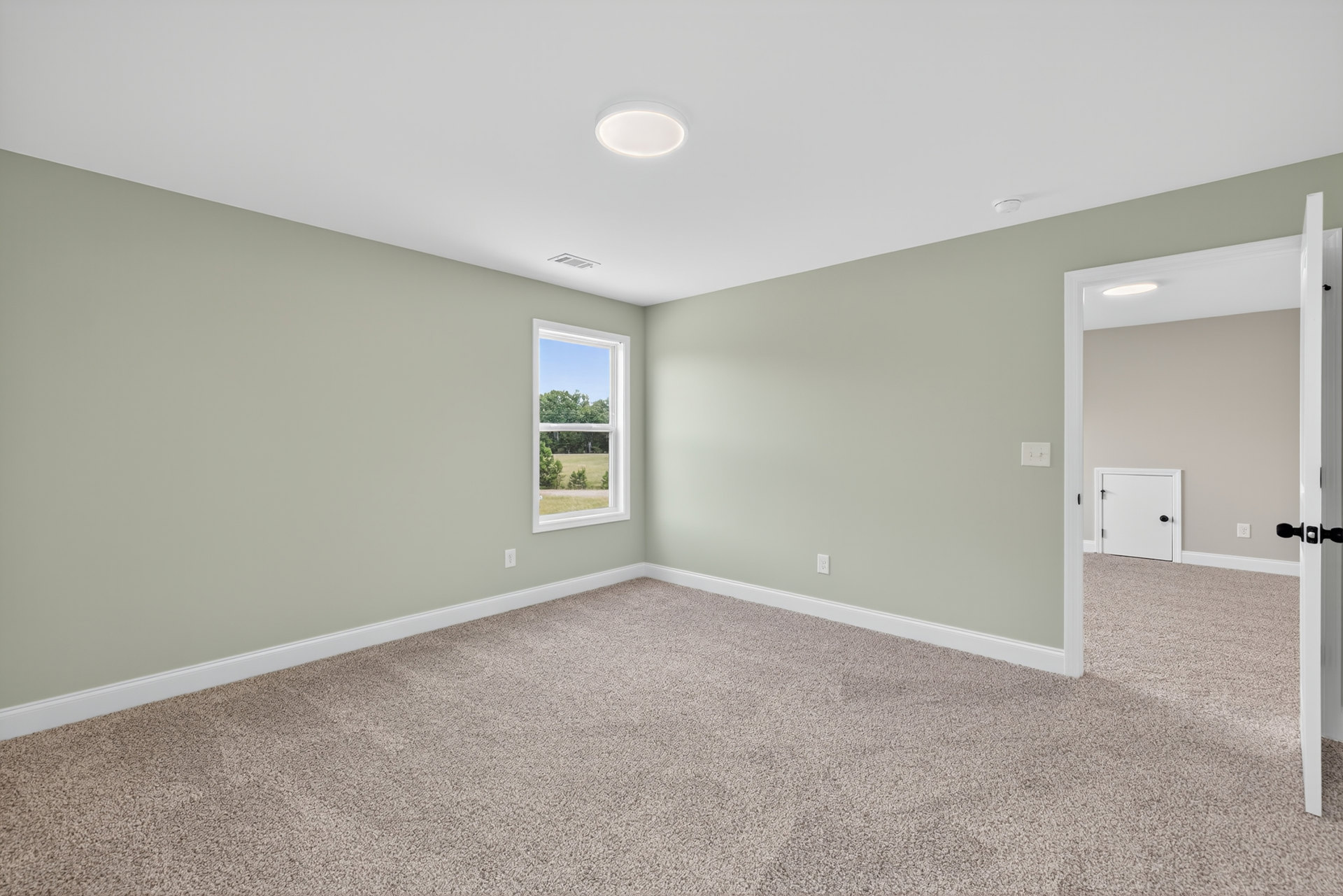 Neutral-toned carpeted floor, white walls, large window with view of green trees, white door with black handle, recessed ceiling light