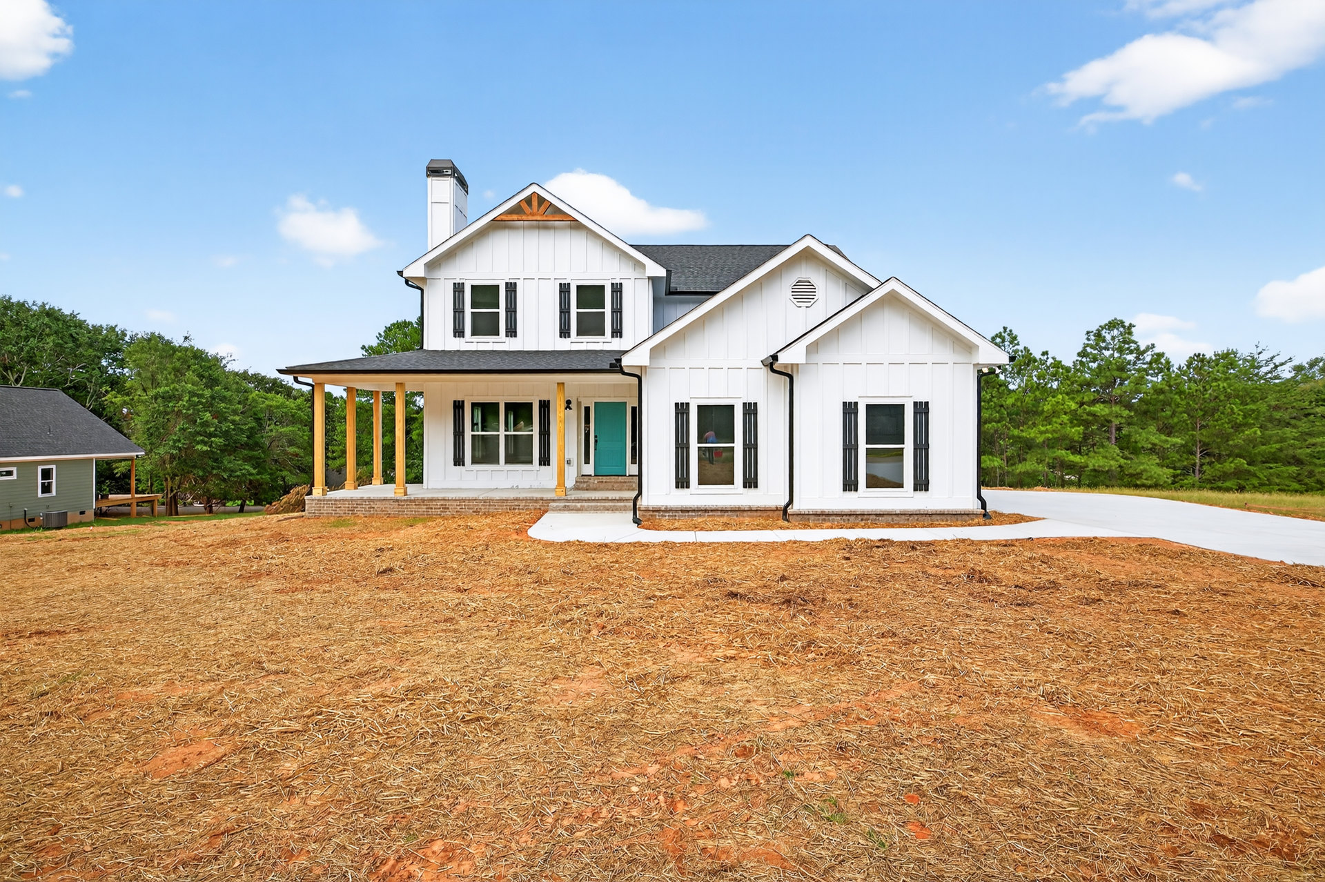 White two-story house with blue front door, black porch railing, white-framed windows, gray shingle roof, spacious green yard, and mature trees under a partly cloudy sky