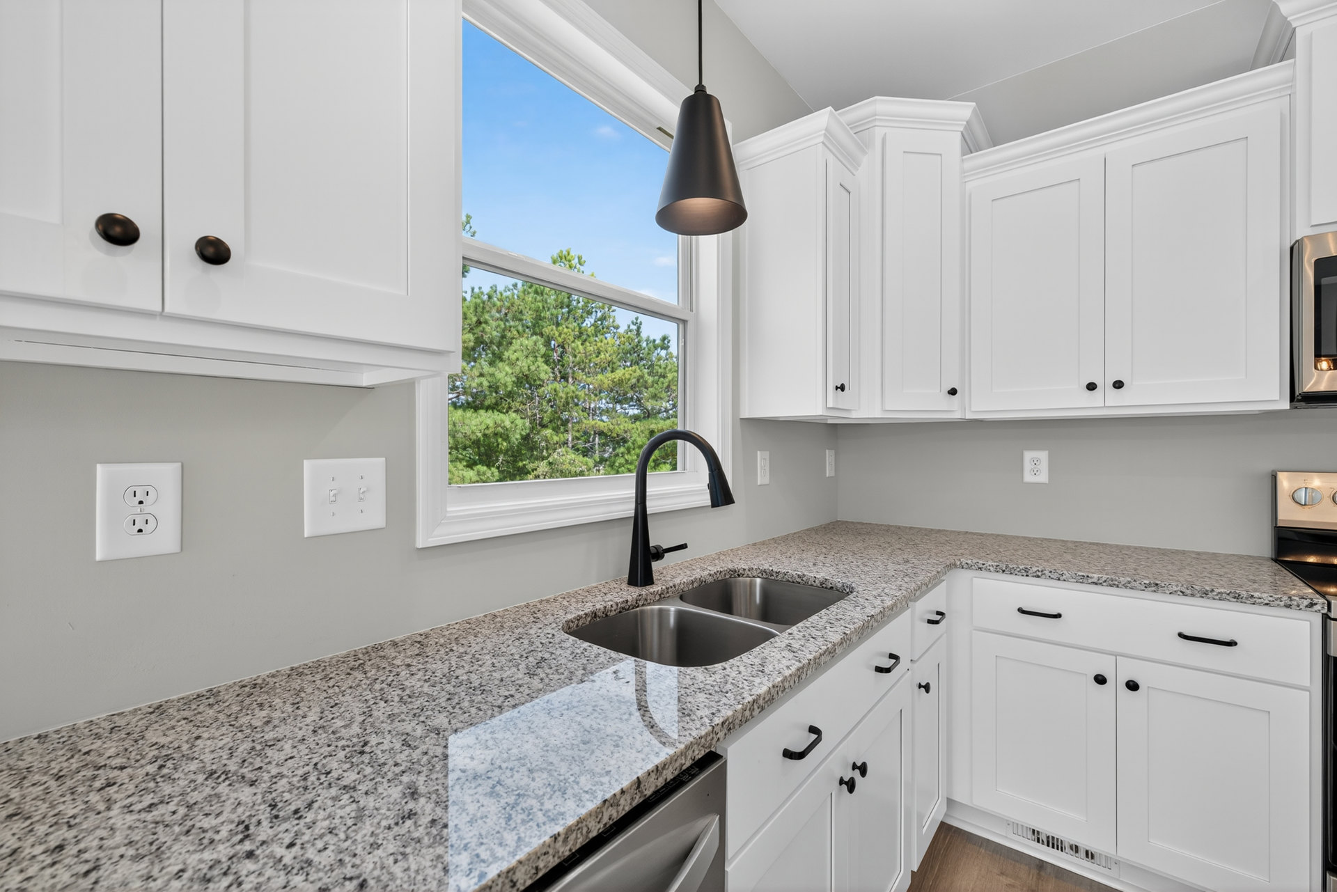 White kitchen with shaker cabinets, stainless steel sink beneath a window overlooking trees, quartz countertop, chrome faucet, black pendant lamp, electrical outlet, and light