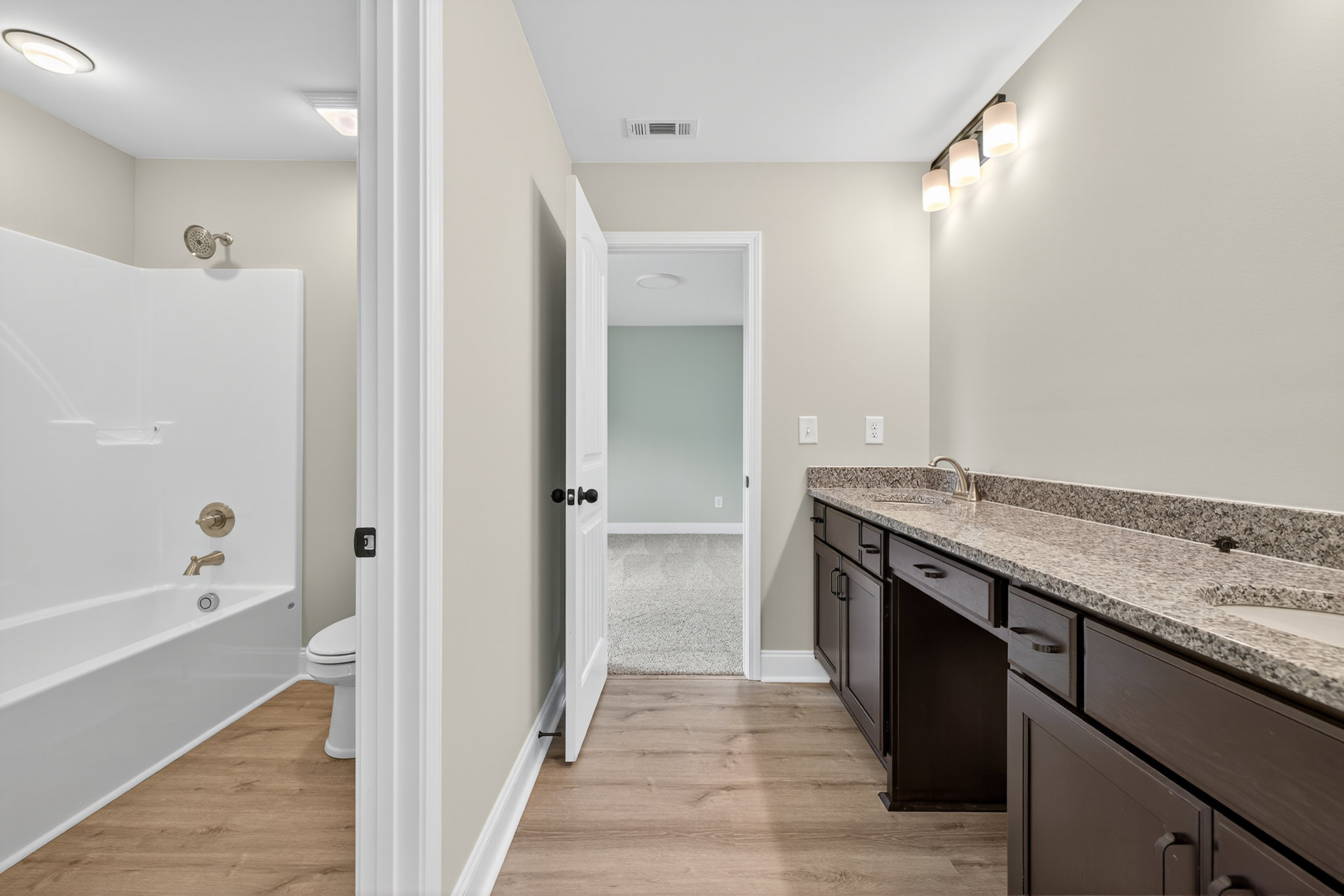 Bathroom with marble countertop, undermount sink, white toilet, tiled floor, bathtub with chrome faucet, open door to adjacent room, three-light fixture, and close-up shower head