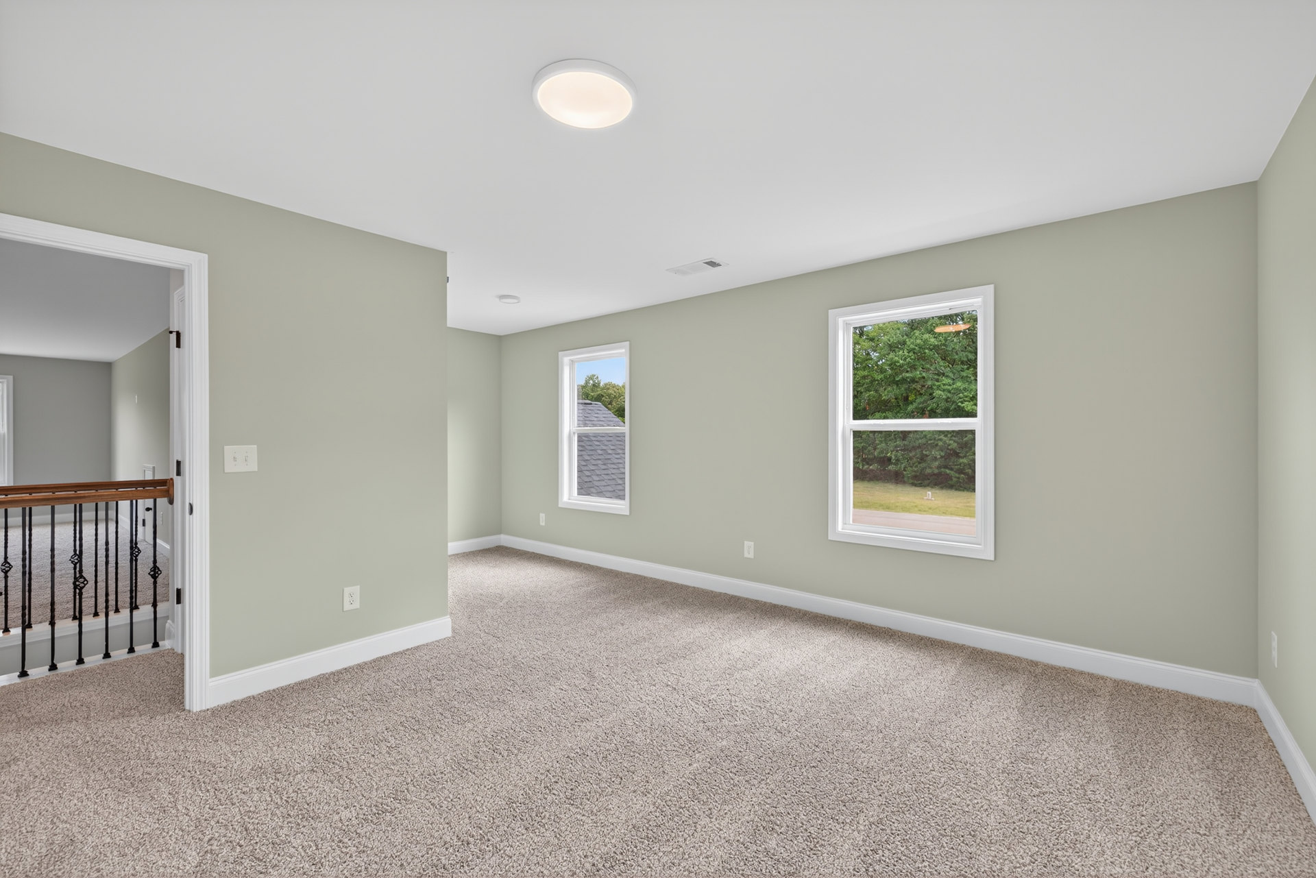 Neutral-toned carpeted room with white-framed windows, painted walls, ceiling light fixture, and close-up of wood railing; view of roof and trees outside.
