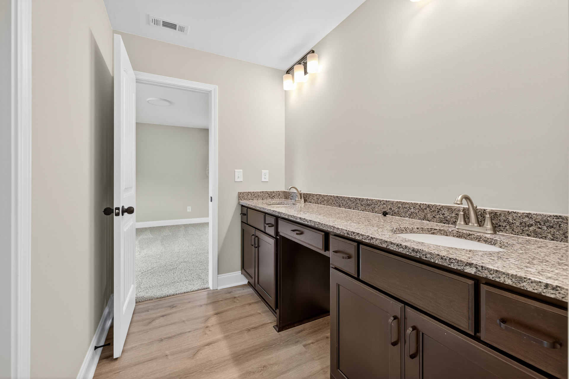 Bathroom with double sinks set in a marble countertop, wood flooring, wall-mounted light fixture, and cabinetry with drawers; faucet and door opening to adjacent room visible.