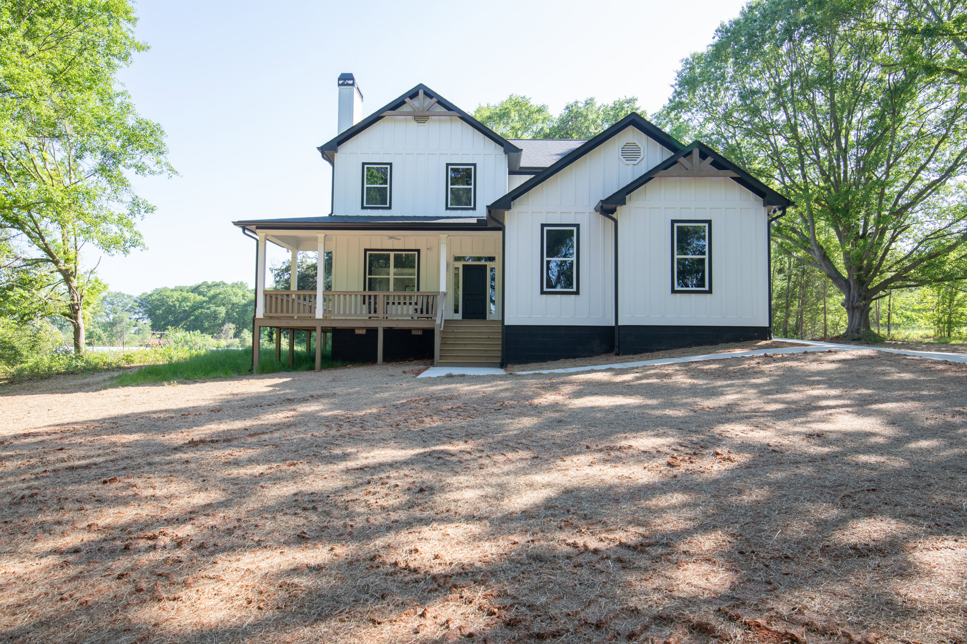 White house with black front door, covered porch supported by white posts, driveway bordered by brown leaves, large windows reflecting trees, overcast sky.