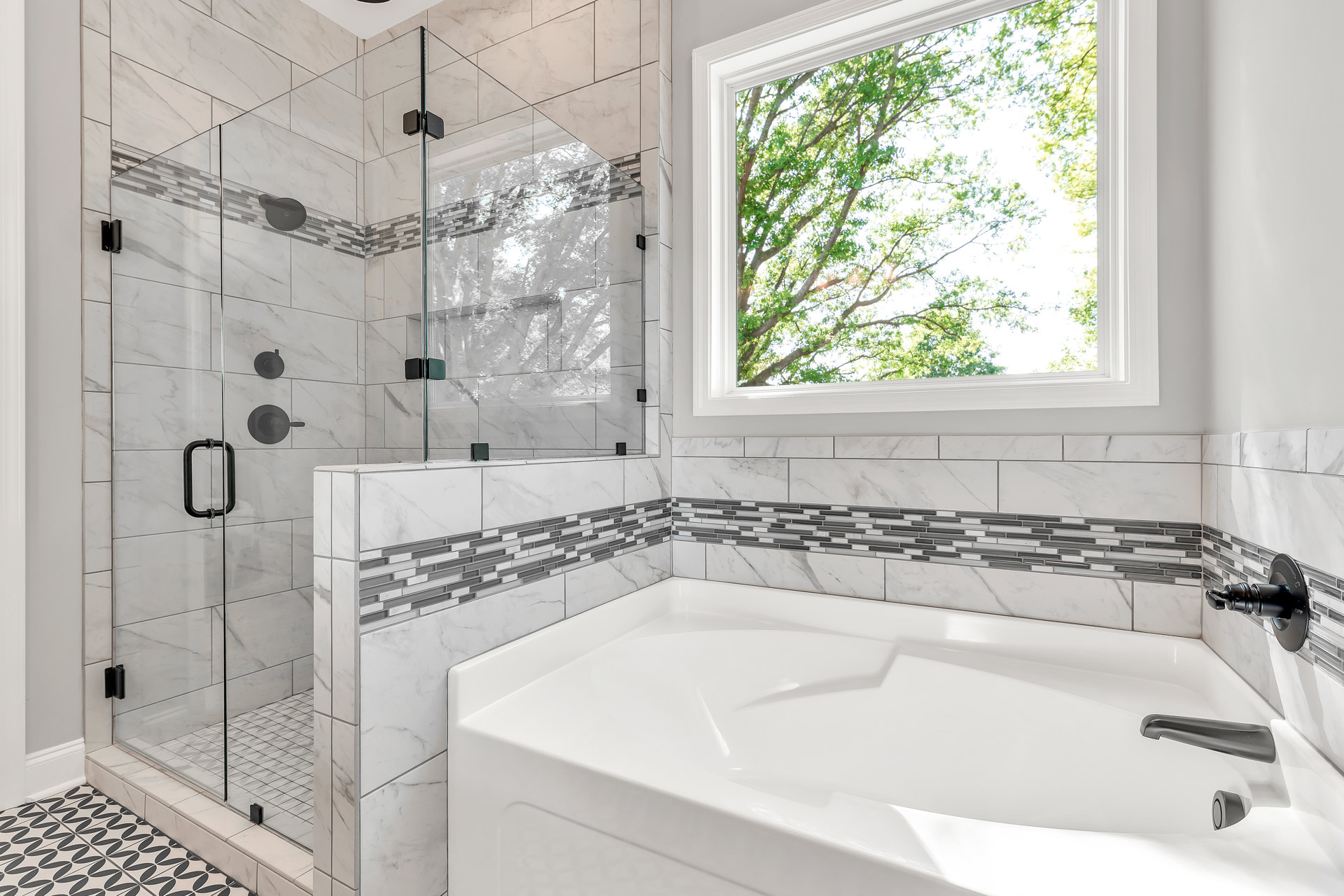 White freestanding bathtub beneath a large window with tree views, patterned tile floor, white tile wall with black handle, modern bathroom fixtures