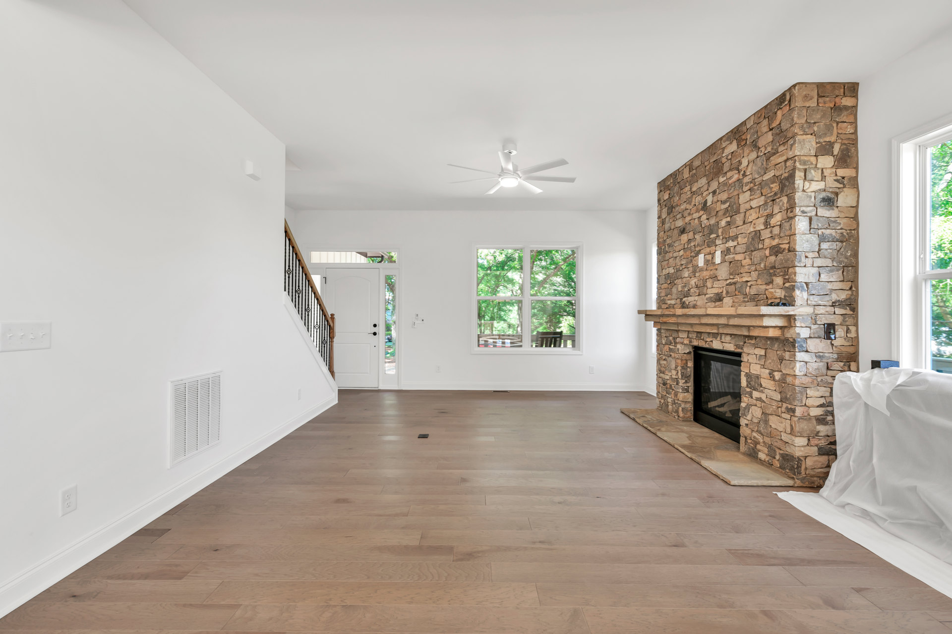 Living room with stone fireplace, hardwood floor, white walls, white ceiling fan with light, and white door