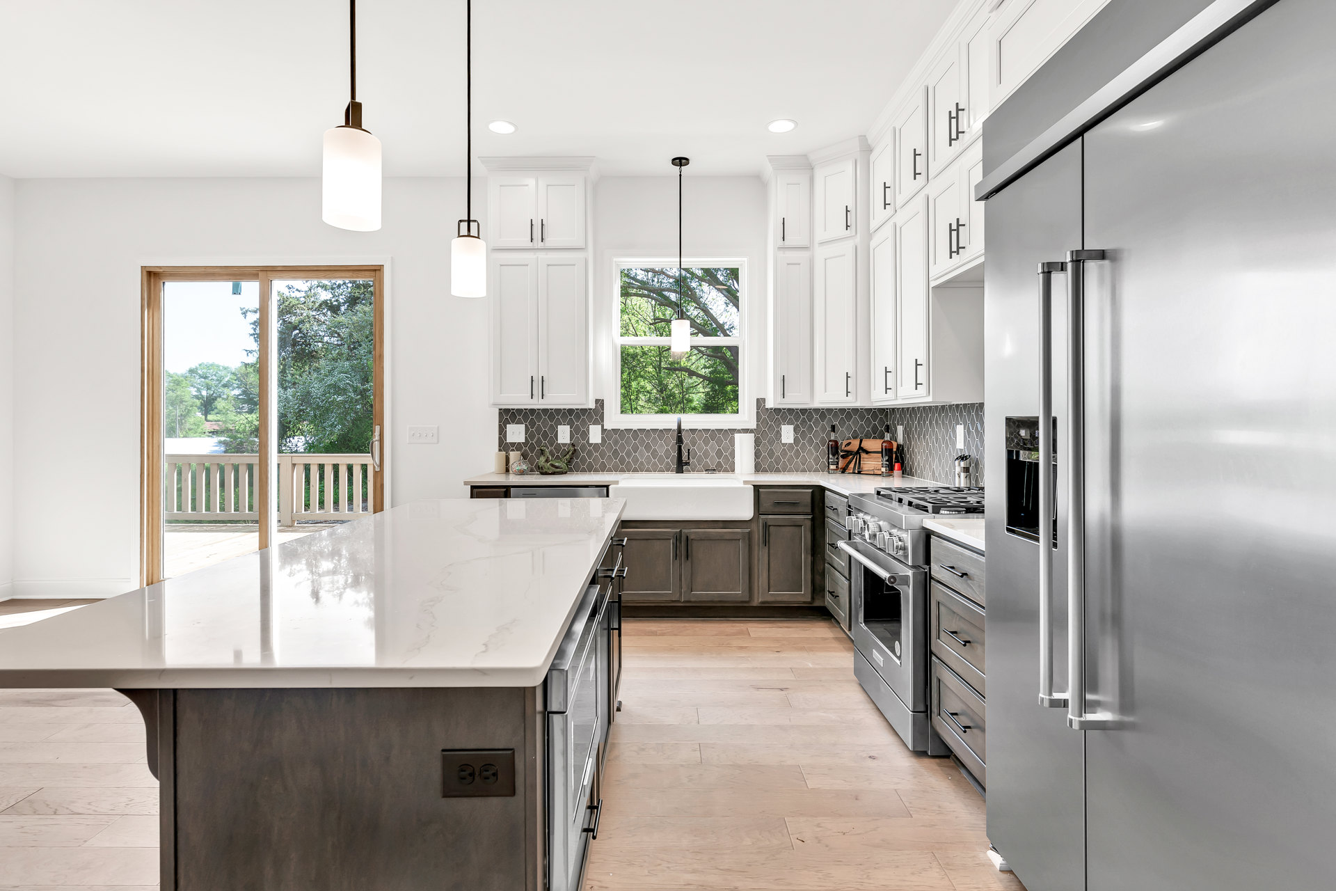 White kitchen with shaker cabinets, large central island with white countertop, stainless steel appliances, window overlooking trees and fence, light wood flooring