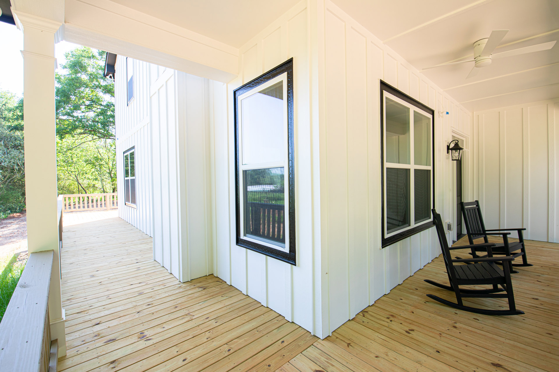 White house exterior with two black rocking chairs on a wooden porch, screened window with white trim, green leafy tree nearby.