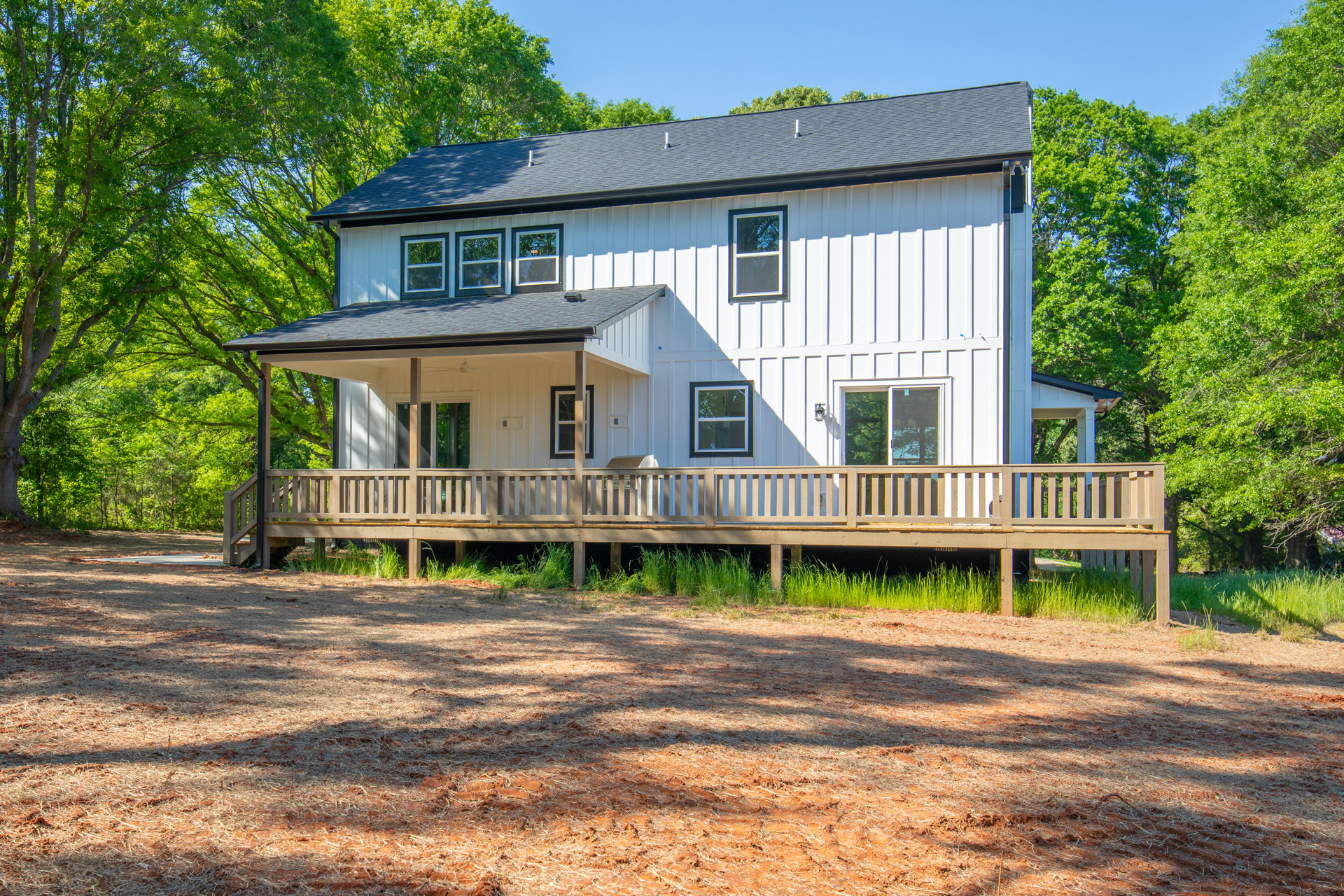 White siding house with covered porch, wooden deck featuring railing, grassy yard, large windows with white frames, mature tree nearby.