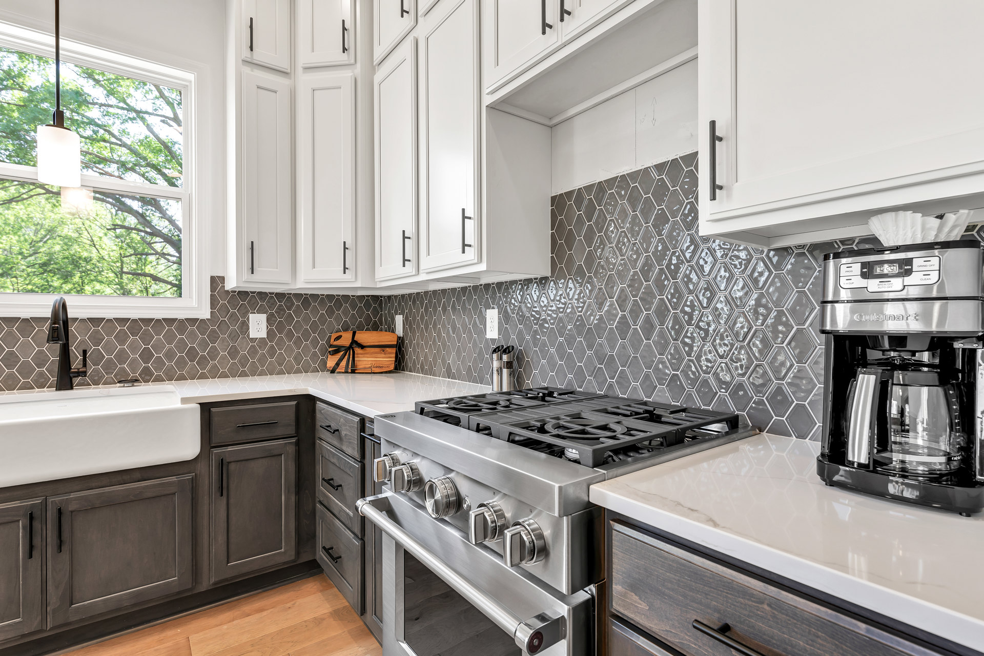 White kitchen cabinets, stainless steel stove, coffee maker with glass carafe, wooden cutting board with black ribbon on countertop, sunlight streaming through window, microwave