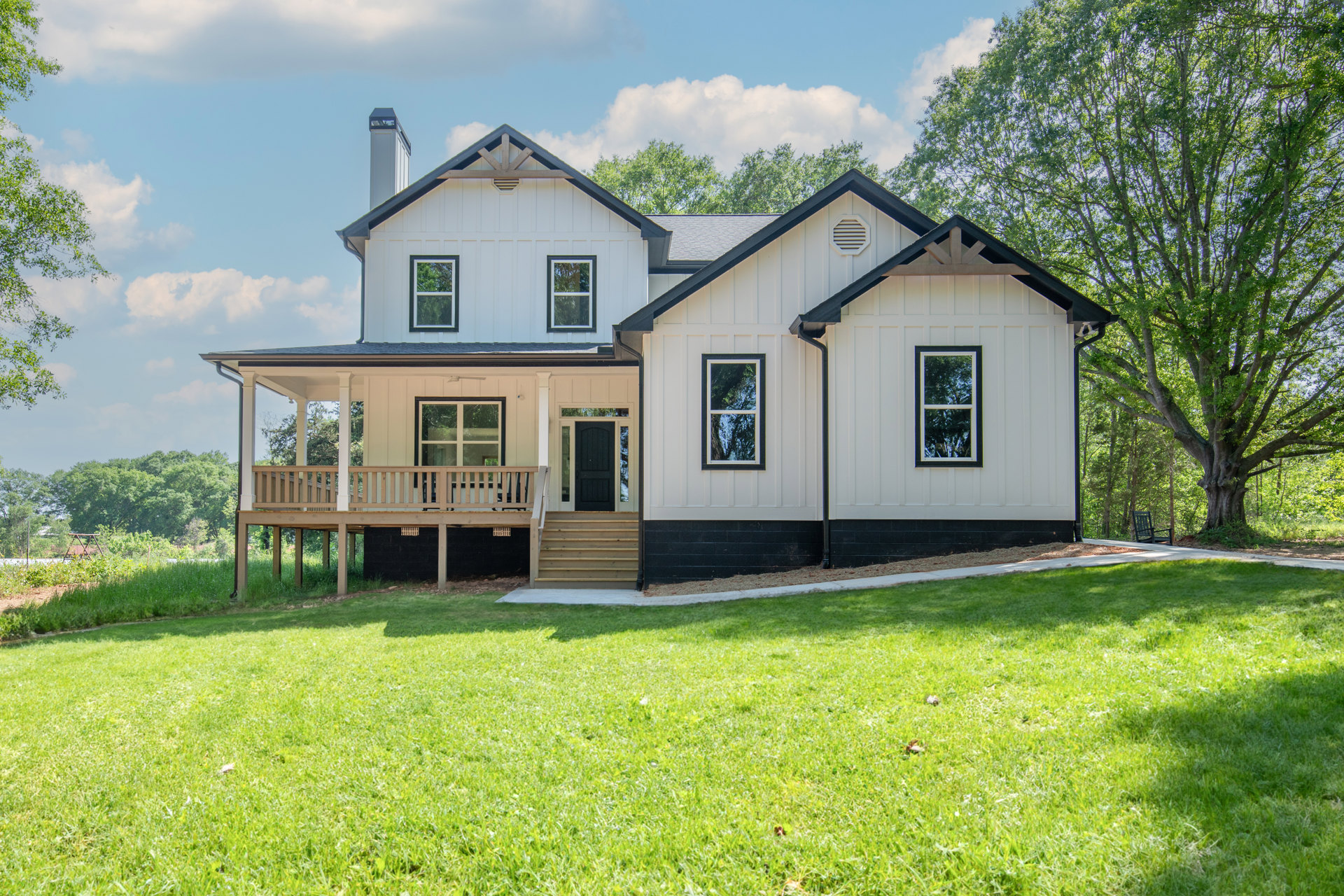 Two-story house with white siding, covered front porch supported by white columns, wooden steps leading to a green lawn, large windows, and mature trees in the background.
