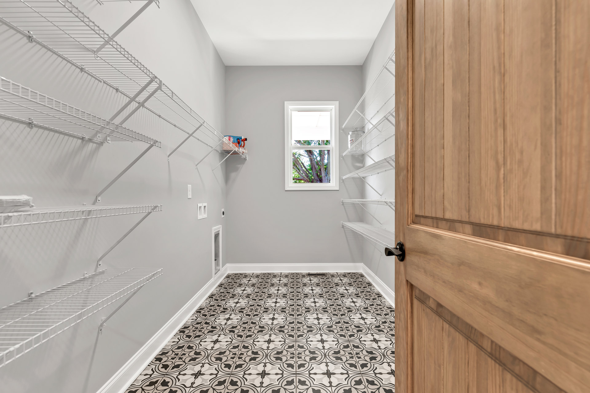 White built-in shelves with laundry detergents, black and white tile flooring, white door, window overlooking trees, plaster walls